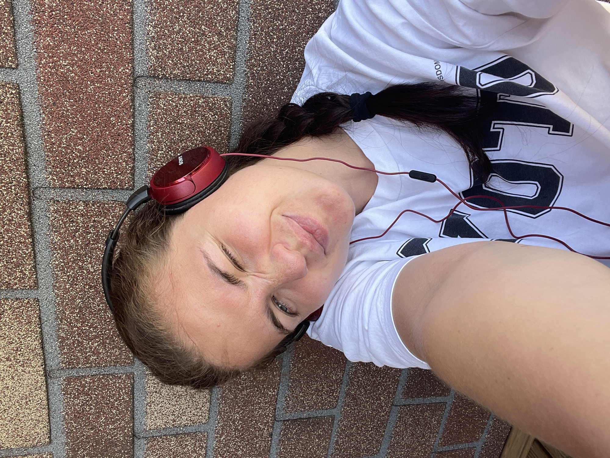 A young woman lies on a paved surface resembling brickwork. She is wearing a white T-shirt with dark graphics, and red headphones. Her dark hair is pulled back in a ponytail. The location is estimated to be in or nearby Мосметаллоконструкция Онт, Russia.