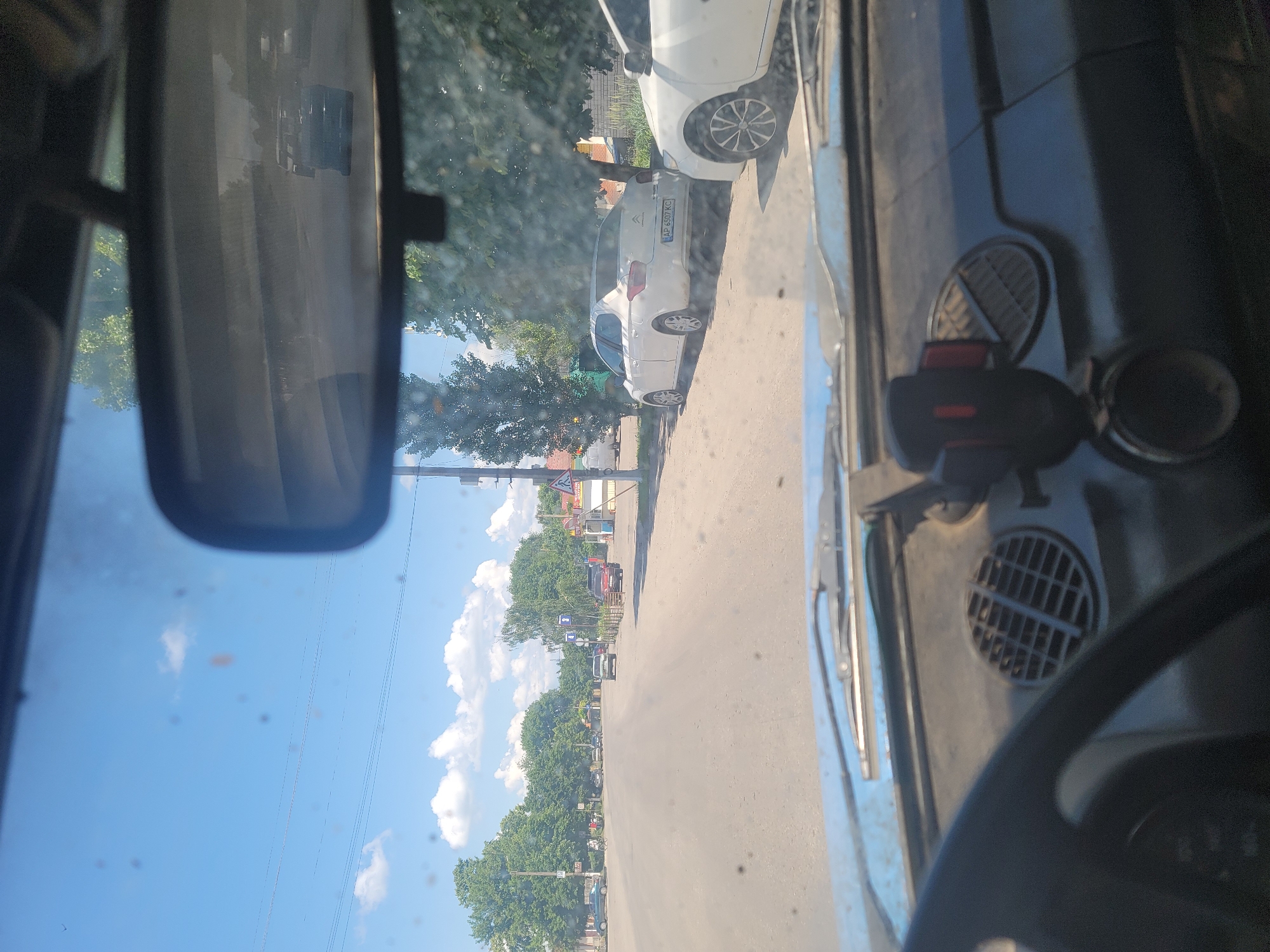 The image is a first-person view from inside a vehicle looking out through the windshield and rearview mirror. The scene appears to be a street in the city of Pereshchepyne, Ukraine, on a sunny day with a blue sky and scattered white clouds. In the foreground, the dashboard of the car is visible, including the steering wheel, a vent, and a phone mount. The road ahead is paved, and several cars are parked along the right side of the road. Trees line the street, and buildings can be seen in the distance. The rearview mirror reflects the road behind the vehicle, showing other cars and the sky. There are no people clearly visible in the image. The environment suggests a typical urban or suburban setting.