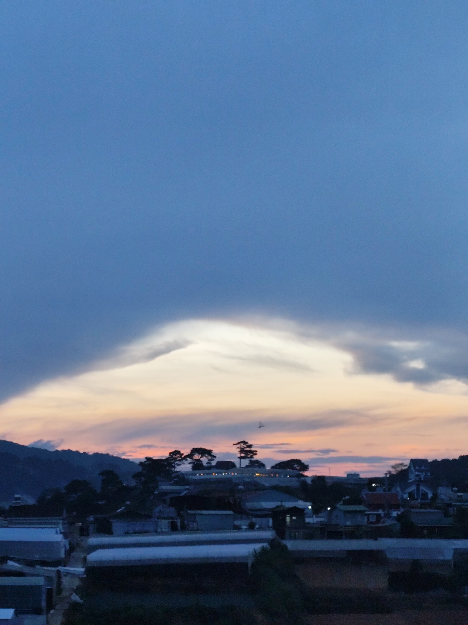 A dusk landscape in Da Lat, Vietnam, shows a view of low-lying structures, possibly greenhouses, in the foreground. Beyond them, silhouetted trees and buildings rise on a gentle hill against a sky with hues of orange and gray. The sky is predominantly covered by a dark, heavy cloud formation with a luminous opening near the horizon. The overall scene is peaceful and muted, with the light indicating either a sunset or sunrise.