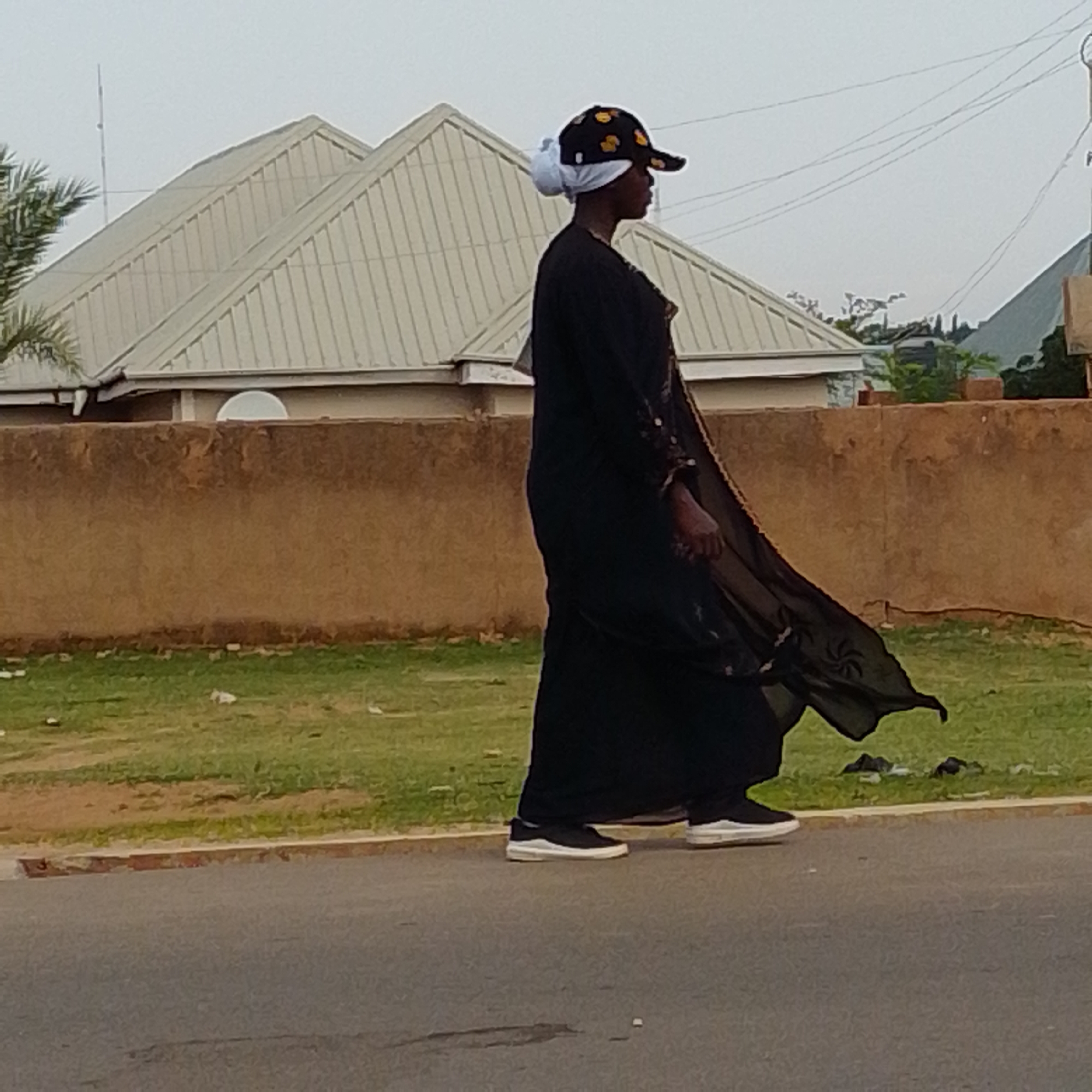 A person walks on a paved road in Gombe, Nigeria, in the side profile view. The person is wearing a black abaya, a white hijab, and a black baseball cap with yellow and black circular patterns. The abaya has a flowing, sheer outer layer with an embroidered floral pattern. The person is also wearing black sneakers with white soles. The background features a brown stucco wall, a patch of dry grass with some litter, a light-colored building with a corrugated metal roof, and some power lines against an overcast sky. The scene appears to be outdoors during the daytime.