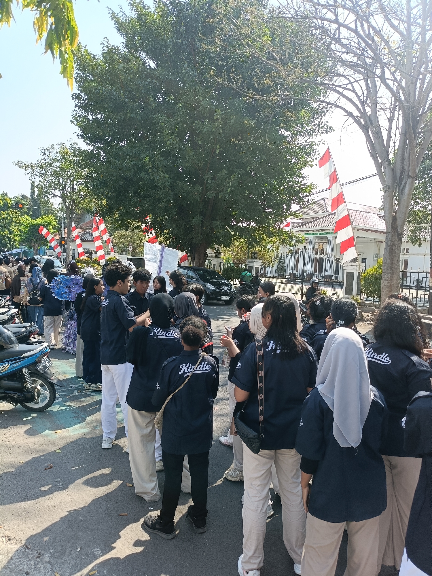The image depicts a group of young people, likely students, gathered outdoors on a sunny day. They are mostly wearing matching dark blue shirts with "Kindle" printed on the back and light-colored pants. Several individuals are wearing headscarves. In the background, there are trees, buildings, and a white and red striped flag which is indicative of the Indonesian flag. Red and white flags are commonly displayed in Indonesia, especially during national holidays or important events.
Specifically, the building in the background with the fence and gate appears to be the Pengadilan Negeri Kota Kediri (Kediri City District Court). The architecture of the building, combined with the presence of the Indonesian flag, strongly suggests this location. The street is paved, and there are motorcycles parked on the side. The overall atmosphere suggests a public gathering or event.