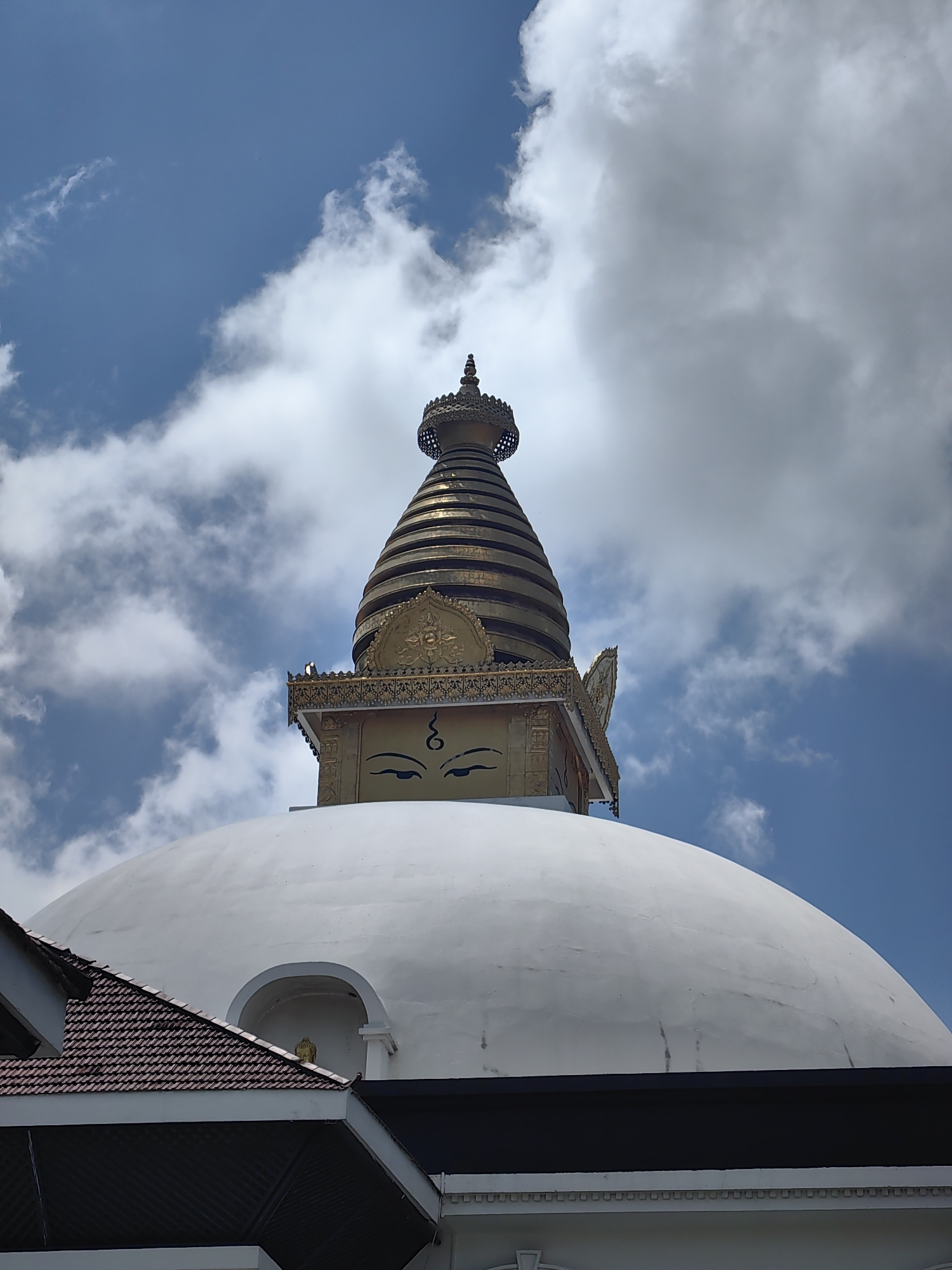 A low-angle medium shot captures the golden spire and upper portion of a Buddhist stupa against a bright blue sky with scattered white and grey clouds. The stupa's conical, tiered golden spire is topped with a decorative finial. Below the spire, a golden cube-shaped structure features two large painted eyes with arched eyebrows, characteristic of Buddhist art. Decorative golden trim adorns the top of this cube. The lower half of the image is dominated by the massive, smooth, white dome of the stupa. Partially visible in the lower left corner is the dark, tiled roof of a building that partially obscures the base of the stupa's dome. The scene appears to be taken outdoors during the daytime, with the sun illuminating the stupa. The overall impression is of a serene and majestic religious monument. This scene is located in Lumbini Bazaar, Nepal, a significant Buddhist pilgrimage site.