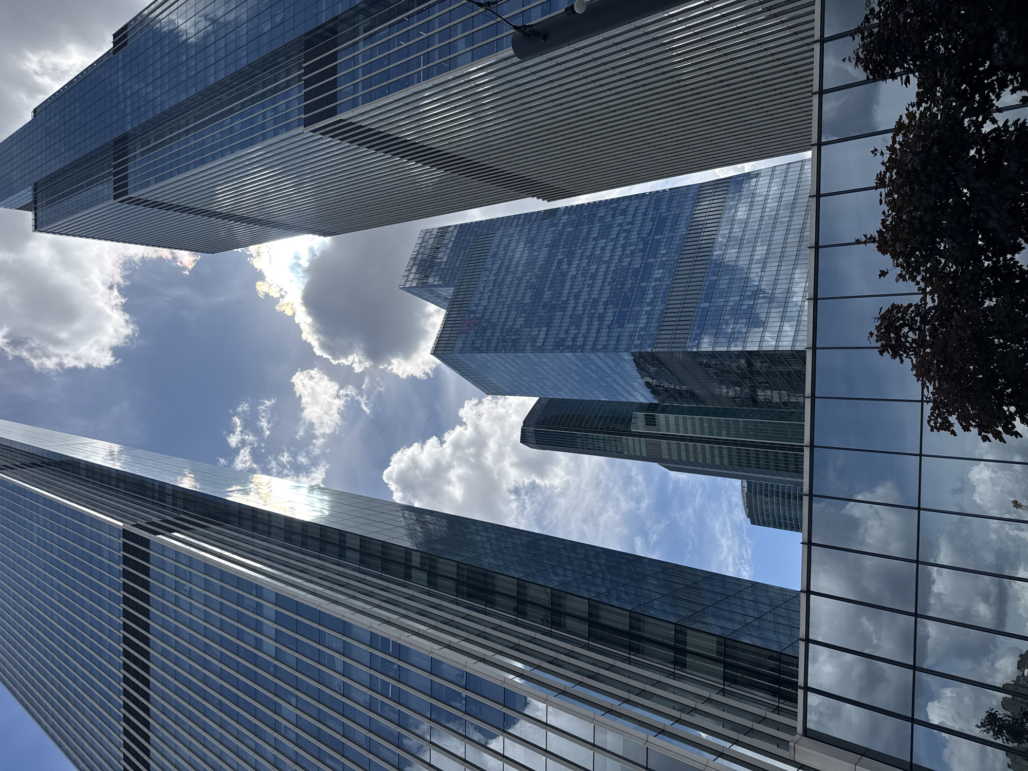 The image captures a low-angle view of modern skyscrapers in Moscow, Russia. The buildings are characterized by their sleek glass facades, reflecting the sky and surrounding clouds. Several buildings are visible, with different architectural styles, including a tall, slender tower with a stepped top and a broader structure with horizontal detailing.
The sky is a mix of blue with scattered white clouds, suggesting it is daytime. Sunlight reflects off the glass surfaces, creating bright highlights on the buildings. A tree with dark foliage is partially visible on the right side of the frame, its reflection also seen in the building's windows.
The perspective is dramatic, emphasizing the height and grandeur of the architecture. The scene conveys a sense of urban modernity and the impressive scale of the city's skyline. There is no visible text in the image. The overall mood is one of awe and the vastness of the urban environment.