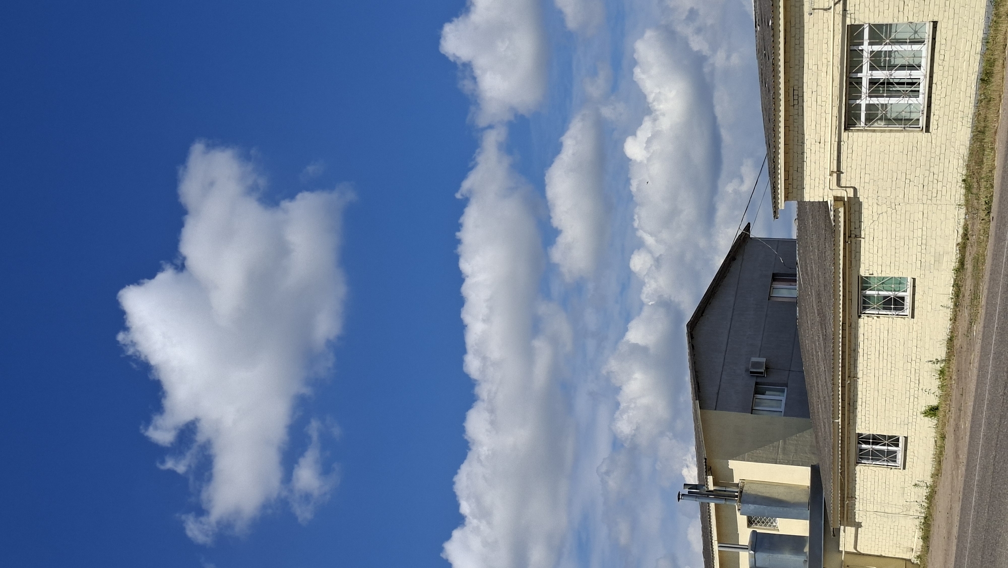 The image depicts an outdoor scene under a bright blue sky with scattered white clouds. In the foreground, a light yellow brick building runs horizontally across the frame. This building has a corrugated metal roof and features several white-framed windows with decorative metal grilles. To the left and behind this building, a taller, multi-story structure is visible, with a section painted yellow and another section in a gray-toned finish. This taller building has a similar corrugated roof and appears to have industrial elements, including large silver cylindrical tanks and vertical exhaust pipes on its left side. An air conditioning unit is mounted on the gray section of the taller building. The scene is located in the area of the Забалоцкі сельскі Савет, Belarus. A paved road runs in front of the yellow brick building, with sparse green vegetation growing along its edge. The overall impression is of an industrial or commercial area on a clear day.