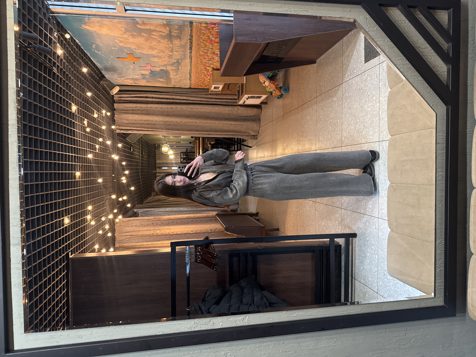 This mirror selfie captures a young woman in an indoor setting in Chaikovsky, Russia. The woman, positioned centrally, is taking a full-body picture using a smartphone, which she holds up to her face. She has long dark hair and is dressed in a grey, textured long-sleeved jumpsuit or loungewear set with dark shoes.
The reflected background reveals a decorative interior space. To the left, a dark grid-patterned wall is strung with numerous warm-toned fairy lights. Above this, a vibrant wall mural depicts a sky with clouds, two stylized airplanes, and a colorful field, possibly of flowers or crops. Below the mural, light brown drapes or curtains are visible. In the mid-ground, a dark wooden console or display unit holds a colorful children's rolling toy or walker. Faint, reversed text, possibly "ADGG" or similar, is reflected on its surface, likely part of a logo. Closer to the foreground, a black clothing rack with multiple hangers holds a folded black jacket or coat. The floor is covered with light-colored, speckled tiles. To the right of the woman, a plush beige upholstered bench or seating area is partially visible. The lighting from the string lights and other fixtures creates a warm and inviting atmosphere.