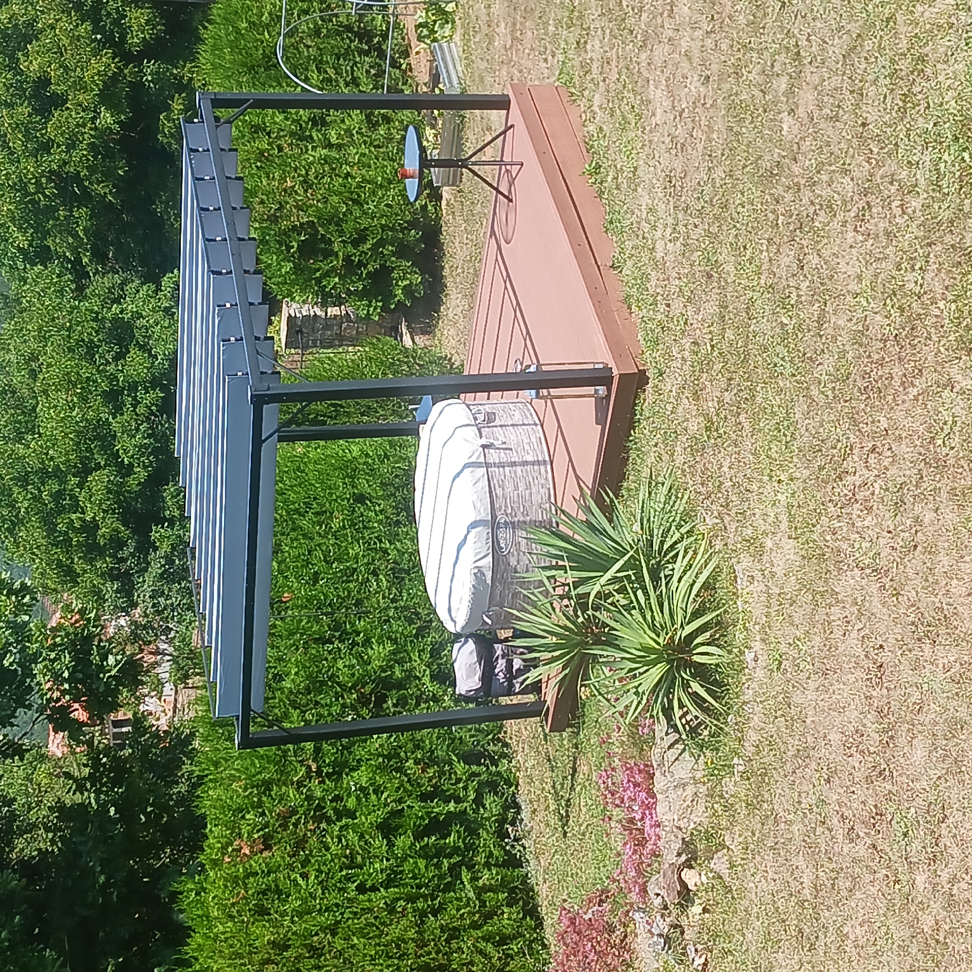 In Sulitsa, Bulgaria, a covered hot tub sits on a wooden deck in a backyard. The hot tub is round and has a grey textured exterior with the "LAY-Z-SPA" logo visible. It is covered with a white, striped tarp. A black metal pergola with a slatted roof covers the hot tub and deck. In the background, there are manicured green hedges and trees, with a stone building visible through the foliage. A small black tripod table with a red object on top stands on the deck to the right of the hot tub. In the foreground, there is dry grass, a Yucca plant, and pink and red flowering plants.