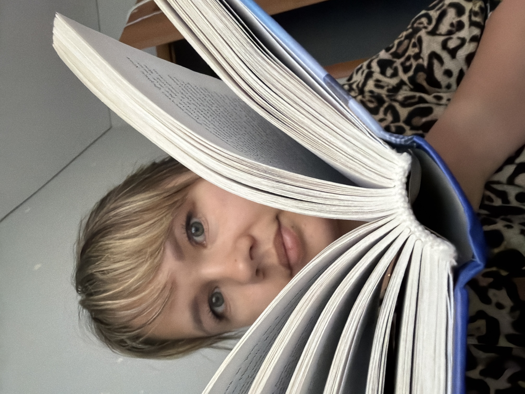 A person is positioned behind a large, open book. The person has light-colored hair, fair skin, and light-colored eyes. The book has a blue cover and numerous white pages filled with text. A pattern resembling animal print is visible behind the book and the person's arm. The photograph is taken in an indoor setting. The location and city details cannot be determined from the image.