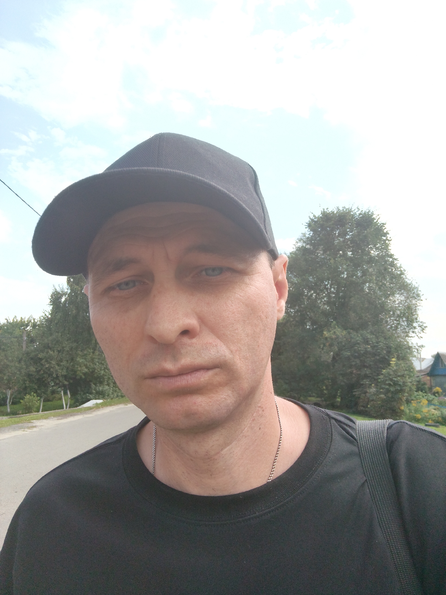 A man wearing a black baseball cap and a black t-shirt is taking a selfie outdoors. He has fair skin, light blue eyes, and his face is visible from the neck up. A silver chain necklace is worn around his neck. The background shows trees, a road, a power line, and houses with visible roofs and windows. The sky is partly cloudy. The location appears to be a rural or suburban area, possibly in Mokshan, Russia, given the architecture of the houses and the general landscape.