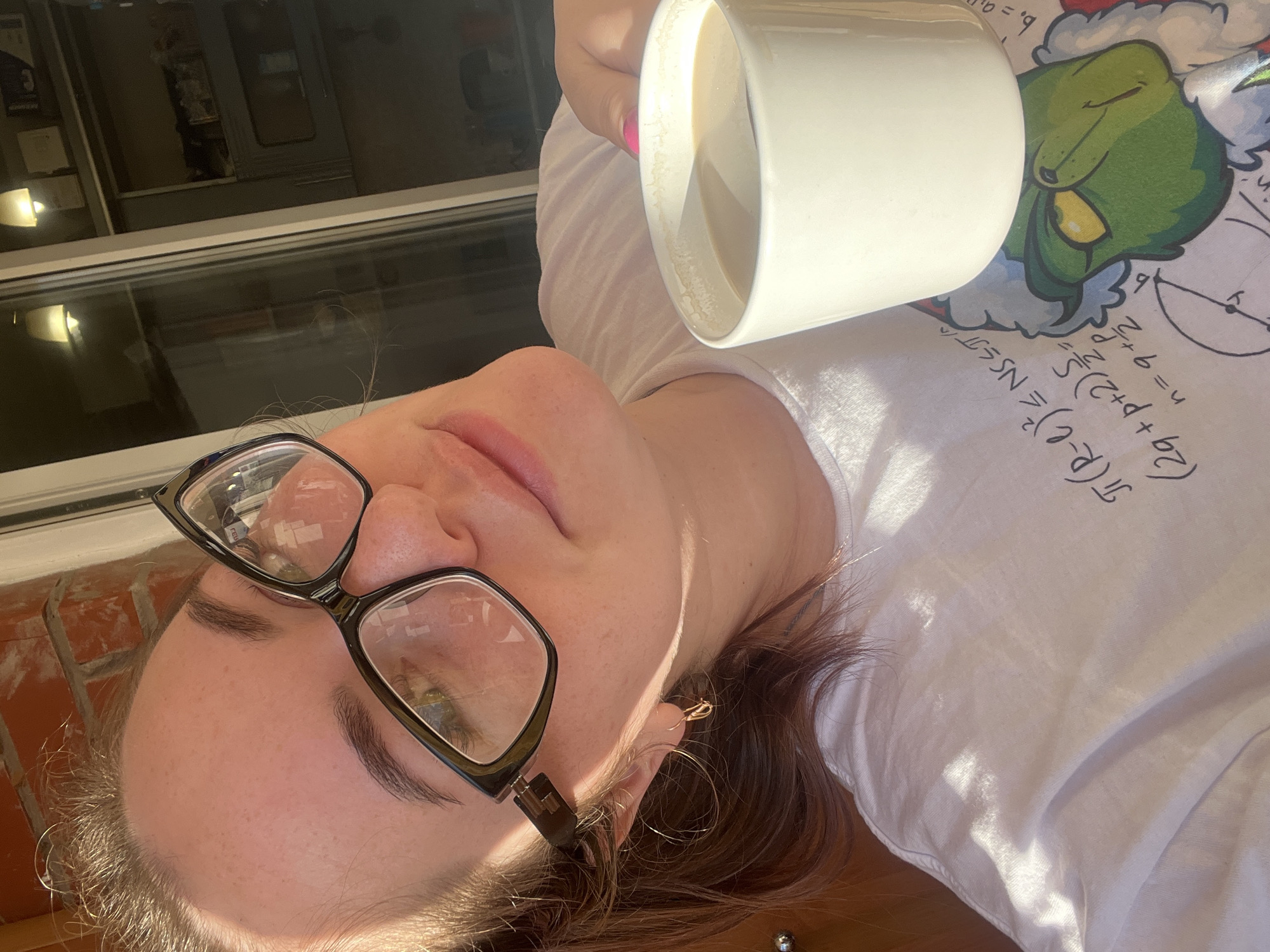 A person with glasses is holding a large white mug in their right hand. The mug appears to contain a light-colored beverage. The person is wearing a white t-shirt with mathematical equations and a cartoon image of the Grinch on it. The background includes what appears to be a window with an outdoor view, along with a brick wall. The location in the city of Chaikovsky, Russia is not identifiable from the image.