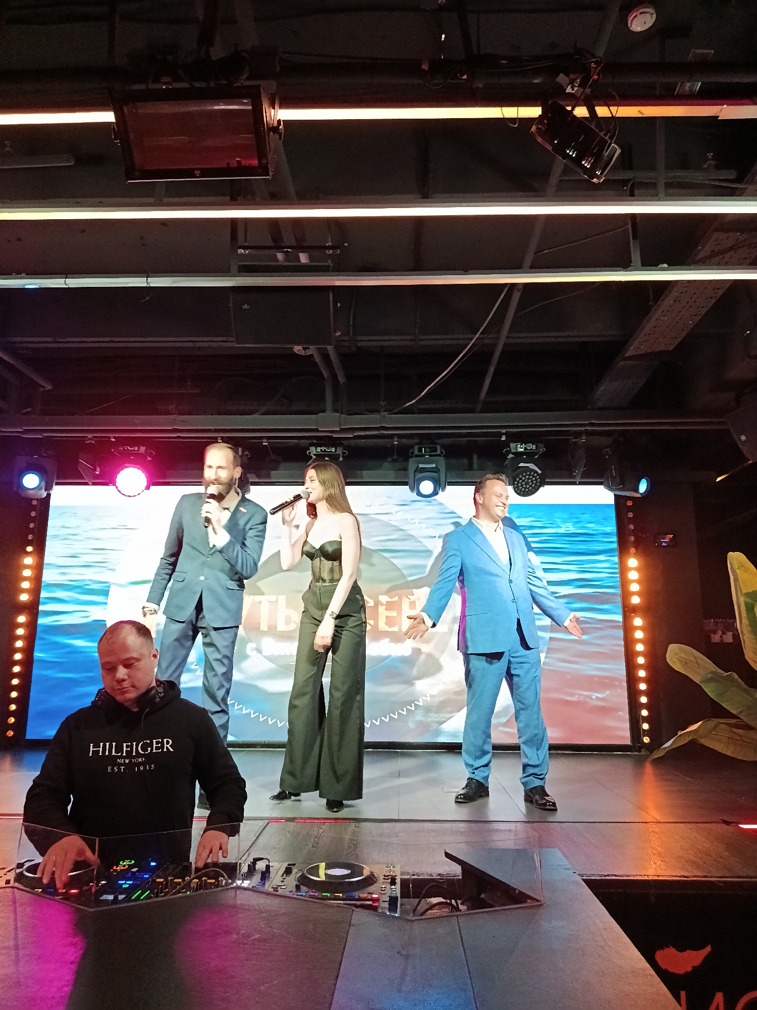 Here is a description of the image:
The image captures a scene on a stage, likely in a club or event venue in Moscow, Russia. Three individuals stand on the stage: two men in suits and a woman in a black corset top and high-waisted trousers. All three are holding microphones, suggesting a performance or presentation. Behind them is a large LED screen displaying what appears to be a water scene. The stage is lit with spotlights.
In the foreground, a man is operating DJ equipment. He is wearing a black hoodie.
The environment includes exposed ceiling beams and lighting fixtures. The overall setting indicates an indoor event with a stage performance aspect.
