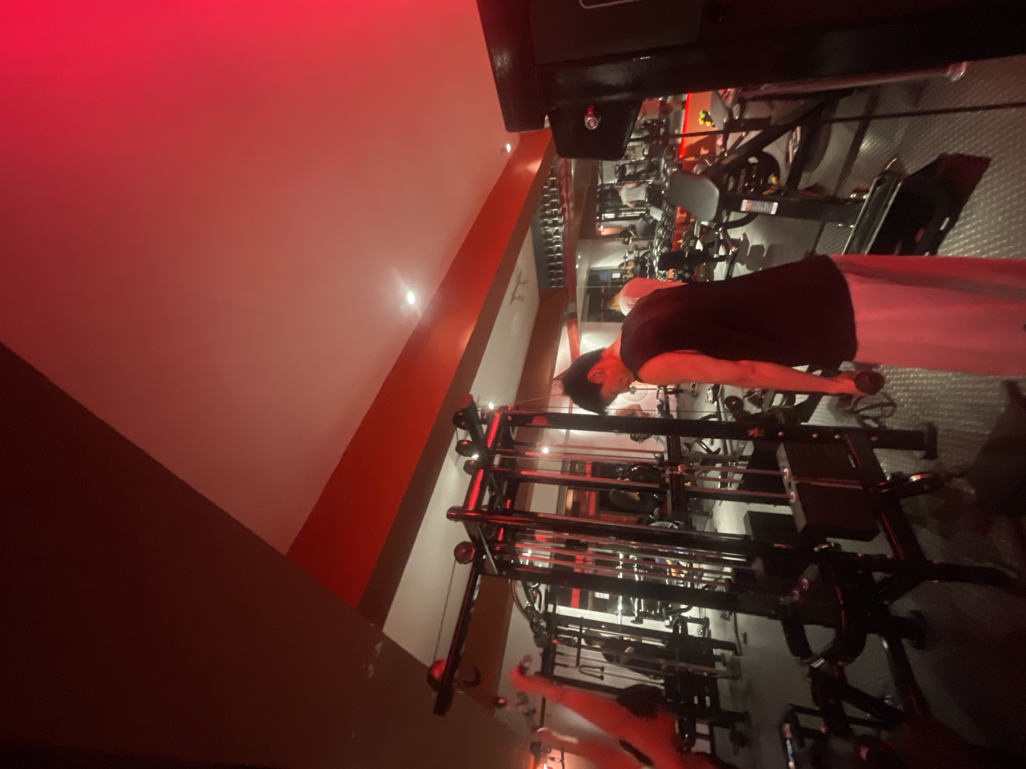 The image captures an indoor gym in the Chongqing High-tech Zone, China, notable for its distinctive red ambient lighting. A young man, dressed in a black tank top and light-colored trousers, is the primary subject, engaged in exercise. He stands, holding a small dumbbell in his right hand, with his arm extended forward, suggesting he is performing an arm or shoulder exercise like a front raise.
The setting is a well-equipped fitness center, featuring various gym machinery including what appears to be a large multi-station cable machine or weight rack system with numerous metal bars and weights. The floor exhibits a patterned texture, possibly rubber gym flooring or diamond plate. The walls are light-colored, contrasting with prominent red horizontal beams or accent strips that run across them, contributing significantly to the red illumination. Small, bright white light fixtures are visible on the ceiling. The overall impression is one of an energetic and focused workout environment. No legible text is visible within the frame.