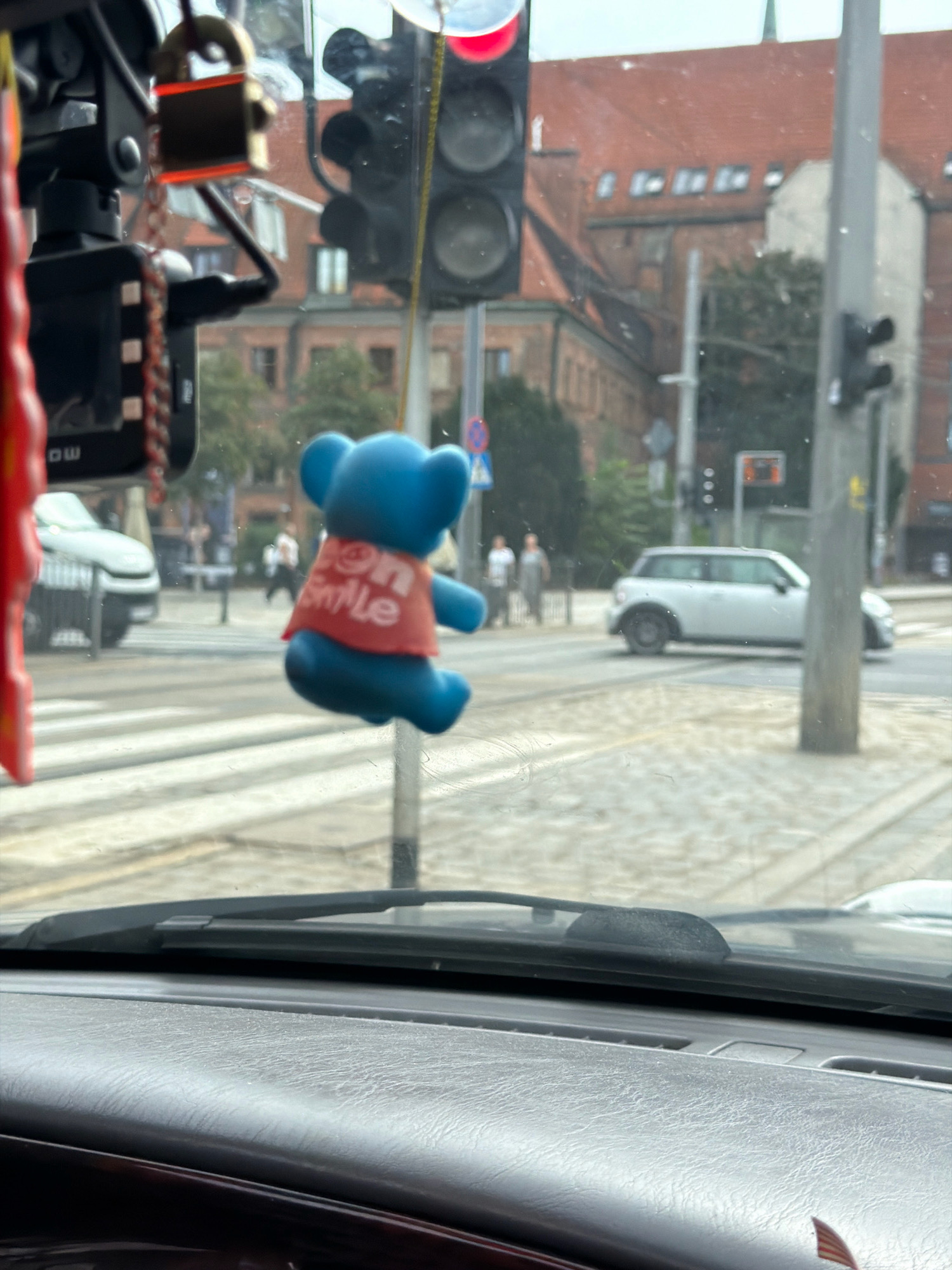 The image captures a view from inside a vehicle in Wrocław, Poland, looking through the windshield. A bright blue teddy bear air freshener, wearing a red shirt with white lettering that reads "On Smile," is prominently suspended from the rearview mirror, obscuring a portion of the traffic lights and building facades. Outside, a white Mini Cooper is visible on the street, driving past a pedestrian crossing. In the background, buildings with red tiled roofs and numerous windows suggest an urban setting. The overall scene conveys a typical city street view, possibly during daylight hours, with a personal touch provided by the car's accessory.