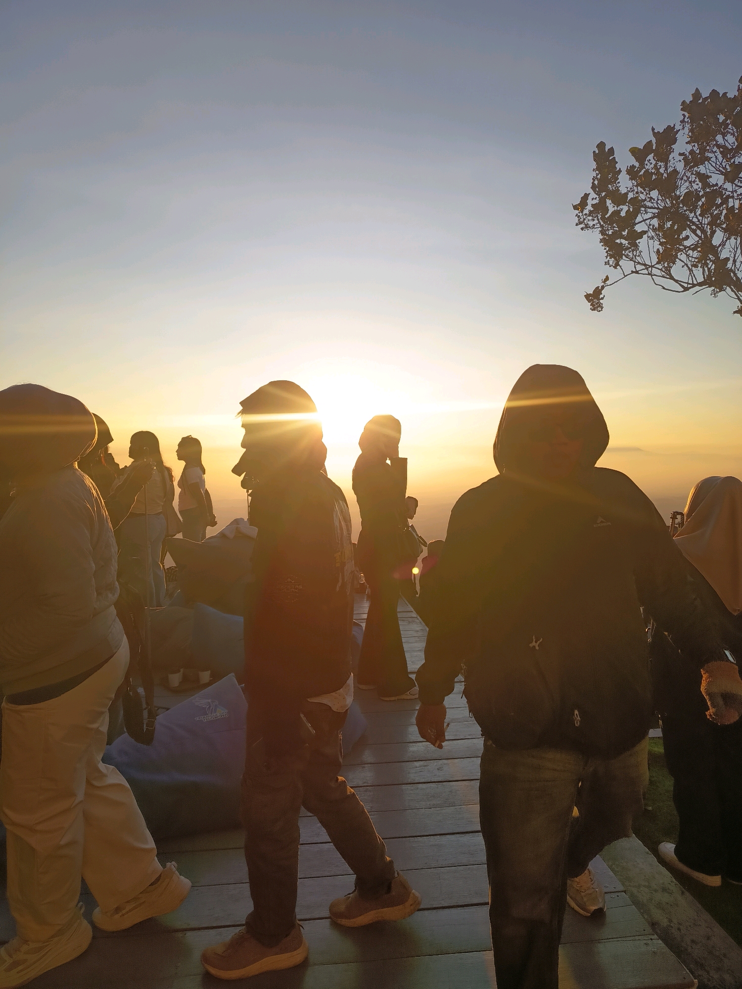 The image depicts a group of people gathered outdoors during what appears to be sunrise or sunset. The sky is a gradient of light blue and yellow, with the sun's rays prominently visible, creating lens flares and silhouetting some of the individuals. The people are standing on a wooden platform. Some are facing the sun, while others are in motion. The environment suggests a natural setting, possibly a viewpoint or hilltop, with a lone tree with dark leaves visible in the upper right corner. The people are dressed casually in jackets, shirts, and pants. One individual in the foreground is wearing a dark hooded jacket and jeans. Another person on the left is wearing a light-colored jacket and pants. There is a blue beanbag or cushion on the wooden platform. The overall atmosphere is that of a shared experience, perhaps a popular tourist spot in Keditan, Indonesia, to witness the dawn or dusk.