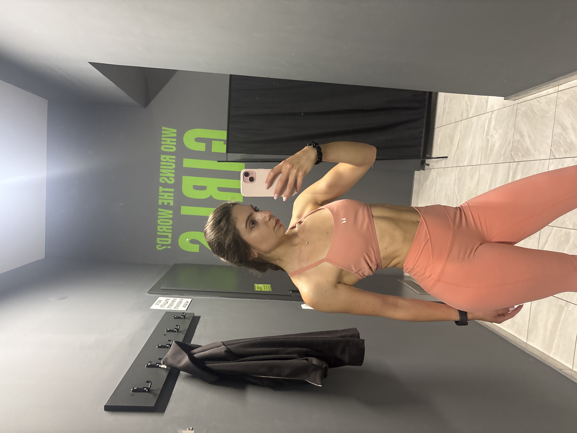 A young woman, likely in her 20s or 30s, is captured taking a mirror selfie in what appears to be a gym changing room or fitness facility in Varna, Bulgaria. She stands sideways, looking into a mirror, holding a light-colored smartphone with her right hand to capture the image. Her left arm is bent, and she wears a black smartwatch on her left wrist and a black scrunchie on her right.
She is dressed in matching athletic wear, consisting of a salmon or light pink sports bra and high-waisted leggings, showcasing a toned physique. A small, delicate necklace is visible around her neck. Her hair is dark brown and styled up, partially visible.
The setting features grey walls, with prominent bright green text painted on one wall. The text is mirrored in the image, but when read forwards, it appears to say "GIRLS WHO RUNS THE WORLD?". A black curtain or divider is seen in the background behind her. The floor is light-colored tiled. On the lower part of the wall, to the left, there is a black coat rack with multiple hooks, on which a dark jacket or coat is hanging. Another small sign with unreadable Cyrillic text is positioned on the wall above the coat rack. The overall impression is a modern and functional fitness-related space.