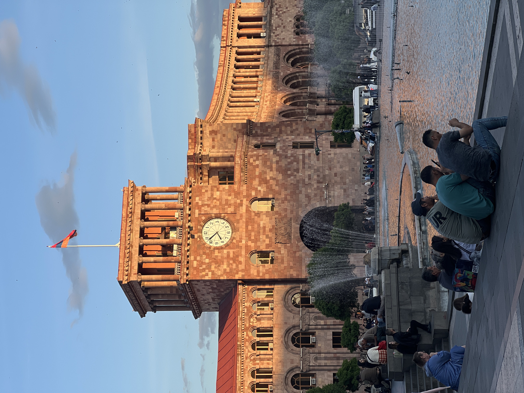 The image depicts a building with a clock tower and an Armenian flag in Unknown, Unknown. A fountain is in the foreground, with several people sitting or standing around it. The architecture is made of red stone with distinct arches. The sky is partly cloudy.