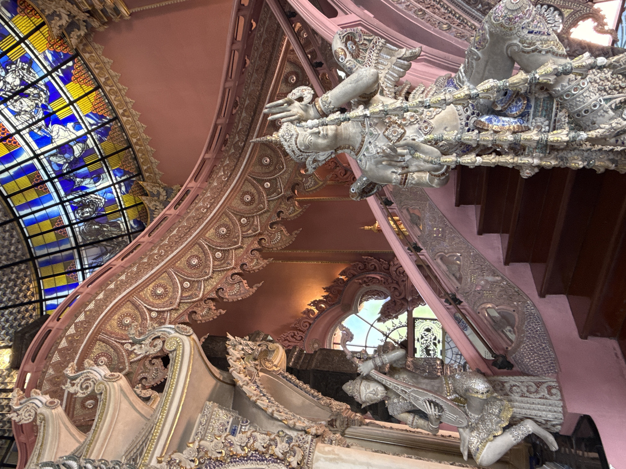 The image captures an interior architectural scene in Samrong Nuea Subdistrict Municipality, Thailand. The focal point is the ornate decoration, featuring statues of human figures with elaborate headdresses and ornamentation. These figures appear to be supporting elements of the structure. A stained-glass window in hues of blue and yellow is visible on the left side, adding color and light to the space. The interior design is characterized by intricate patterns and detail work. A pinkish-brown color palette dominates the walls and ceiling. A staircase with dark wooden steps is visible on the right side. The overall impression is one of elaborate decoration within a cultural and possibly religious context.