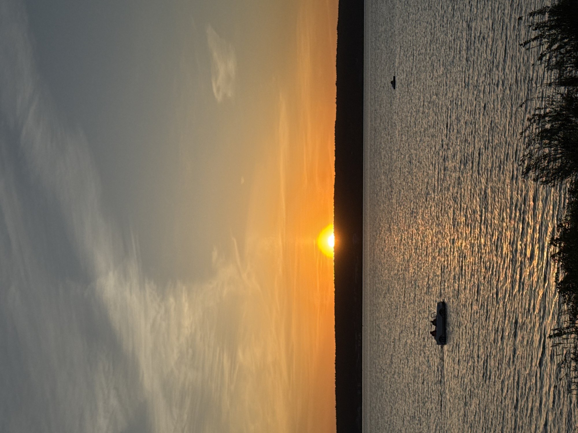 The image depicts a sunset over a body of water. The sun is positioned on the horizon, casting a golden glow across the sky and reflecting on the water's surface. Two boats are visible on the water, one closer to the viewer and the other further in the distance. Trees line the edge of the water on the right side of the image. The sky is a mix of light gray and yellow hues, with wispy clouds scattered throughout.