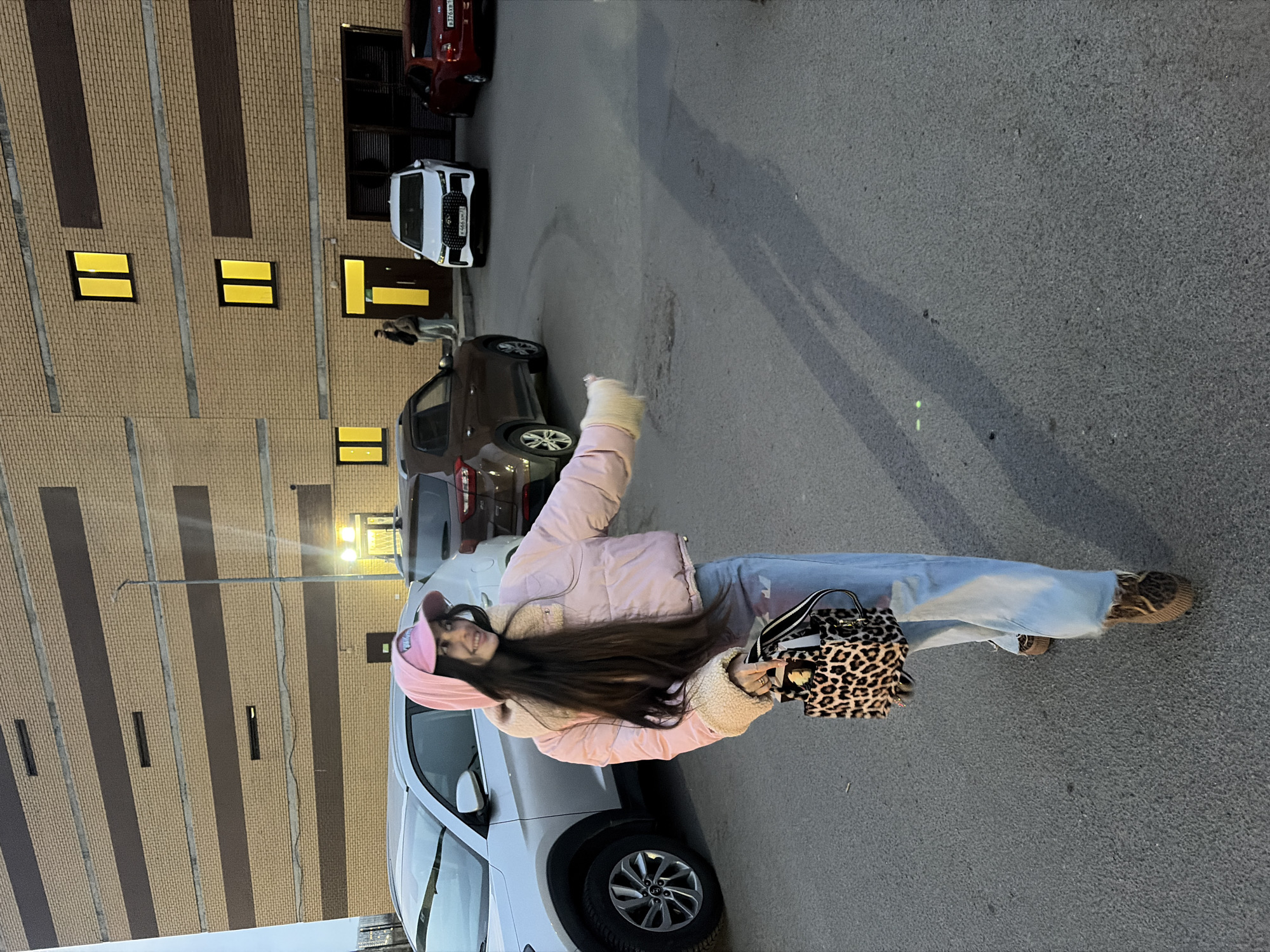 A young woman stands in a parking lot beside a silver car, holding a leopard-print purse. She wears a pink hat and jacket, with light-blue jeans, and has long dark hair. The setting appears to be Saint Petersburg, Russia, based on the urban architecture of a tall building with many windows and the presence of other cars. The ground is asphalt, and it is likely dusk or early evening based on the lighting.