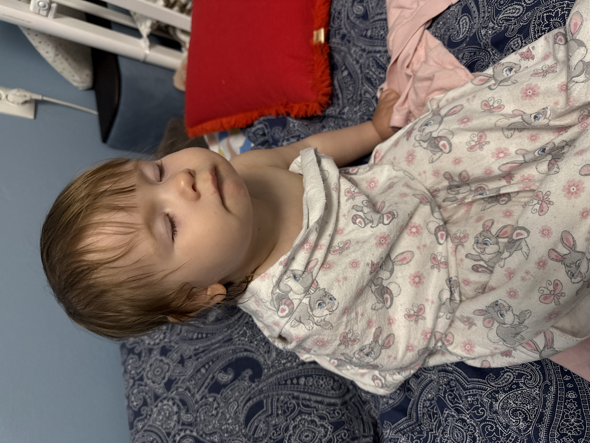 A child is lying on a bed with blue paisley patterned bedding. The child is covered in a white blanket patterned with grey rabbits and pink flowers. The child has light brown hair and closed eyes. A red pillow with a fringed edge is visible near the top of the frame. A partially visible baby crib is in the background. The scene does not depict the location in the city of Moscow, Russia.