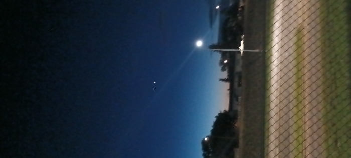The image captures an outdoor twilight or nighttime scene within the city of Medicine Hat, Canada. The upper sky displays a dark blue hue, fading to a lighter blue with a subtle orange-pink gradient near the horizon. Two distinct bright, non-point sources of light are prominent in the upper-mid left quadrant of the sky. A faint, linear light trail is visible below these objects. In the midground, silhouetted trees and indistinct building structures are discernible against the low light of the horizon. A tall, upright street lamp with an active, bright white light fixture is positioned on the right side of the frame, casting a visible light beam diagonally towards the left. The lower third of the image is occupied by a chain-link fence, spanning across the foreground from left to right. Beyond the fence, a flat expanse of ground, appearing as a field or open area, contains a lighter-toned pathway or track. No individuals, specific actions, or interactions are depicted within the scene.