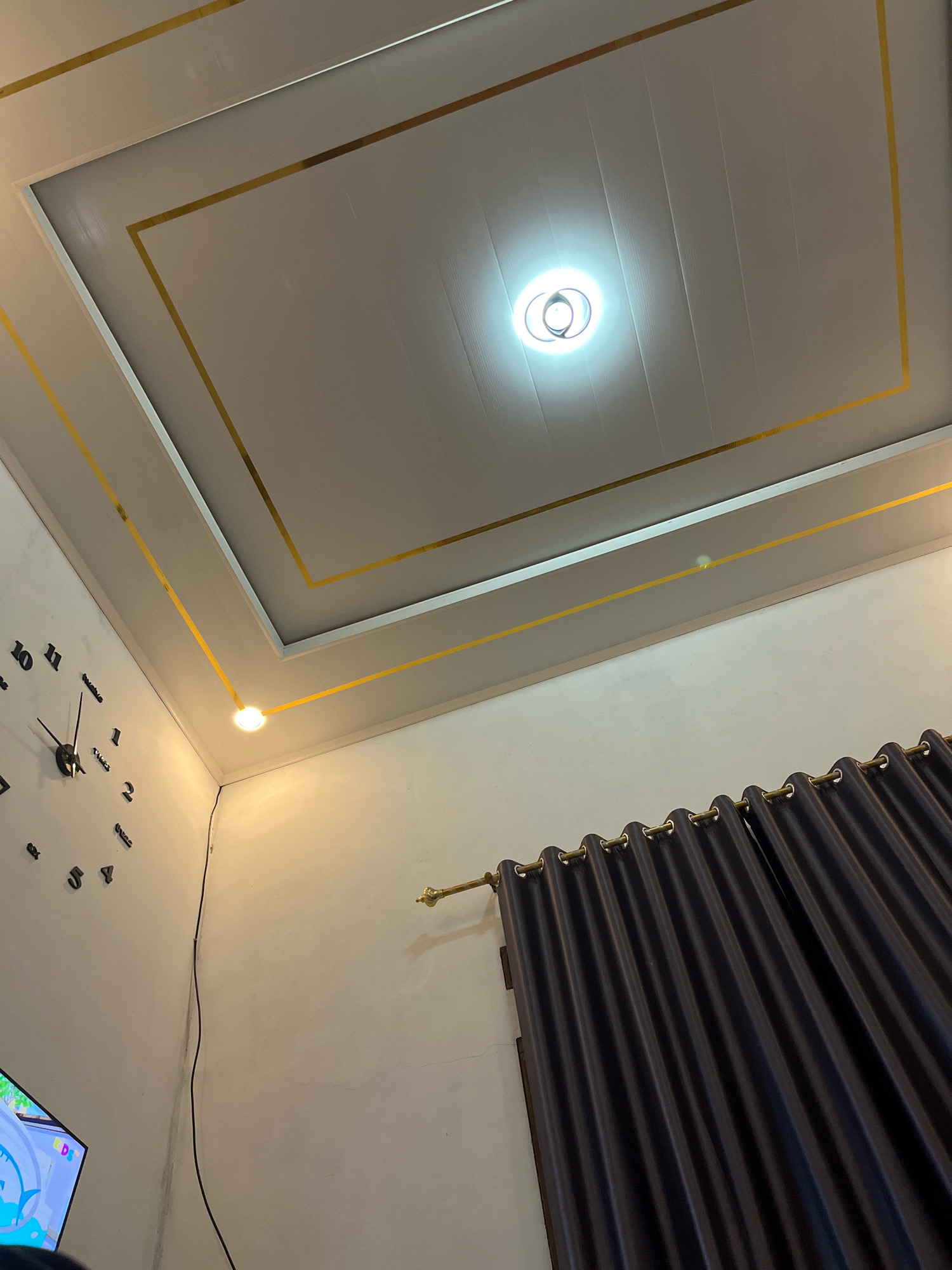 The image depicts the interior of a room, likely a living space, in Kebumen, Indonesia. The focus is on the ceiling, which is a suspended white ceiling with a decorative rectangular pattern outlined in gold trim. A circular ceiling light fixture is centrally located within the pattern, emitting a bright glow. To the left, a large, modern wall clock with black numbers and hands is mounted on a white wall. Below the clock, a portion of a television screen is visible, displaying colorful graphics. In the lower right of the image, dark gray curtains with gold grommets hang from a curtain rod. A black electrical cord runs vertically down the wall from the ceiling near the clock. The lighting suggests it is daytime or evening, with the ceiling lights providing illumination. The scene appears to be a domestic setting, neatly arranged.