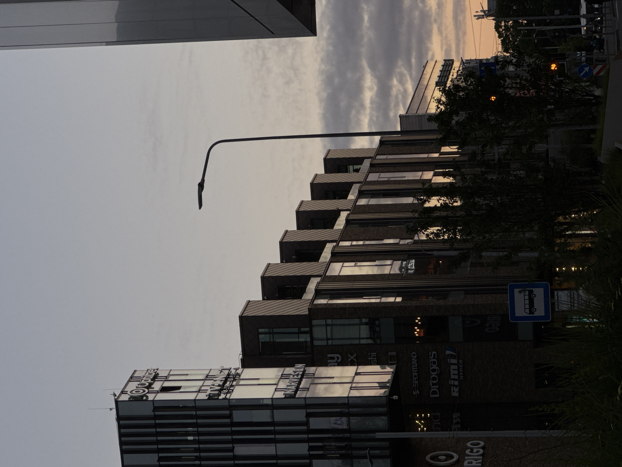 A low-angle, full shot captures the facade of the Origo shopping center in Riga, Latvia, at dusk. The modern building, constructed with glass and dark brick, features multiple levels with visible signage for stores such as "SPORTLAND", "Drogas", and "Rimi HYPER". A distinctive tall, slender streetlight with a curved arm extends upwards on the right side of the frame. The sky above is a muted, hazy gray, with streaks of pale orange near the horizon, indicating the sunset. Several trees with dark foliage are situated in the foreground, partially obscuring the lower levels of the building. A blue bus stop sign with a white bus icon is visible. In the distance, additional buildings and streetlights can be seen.