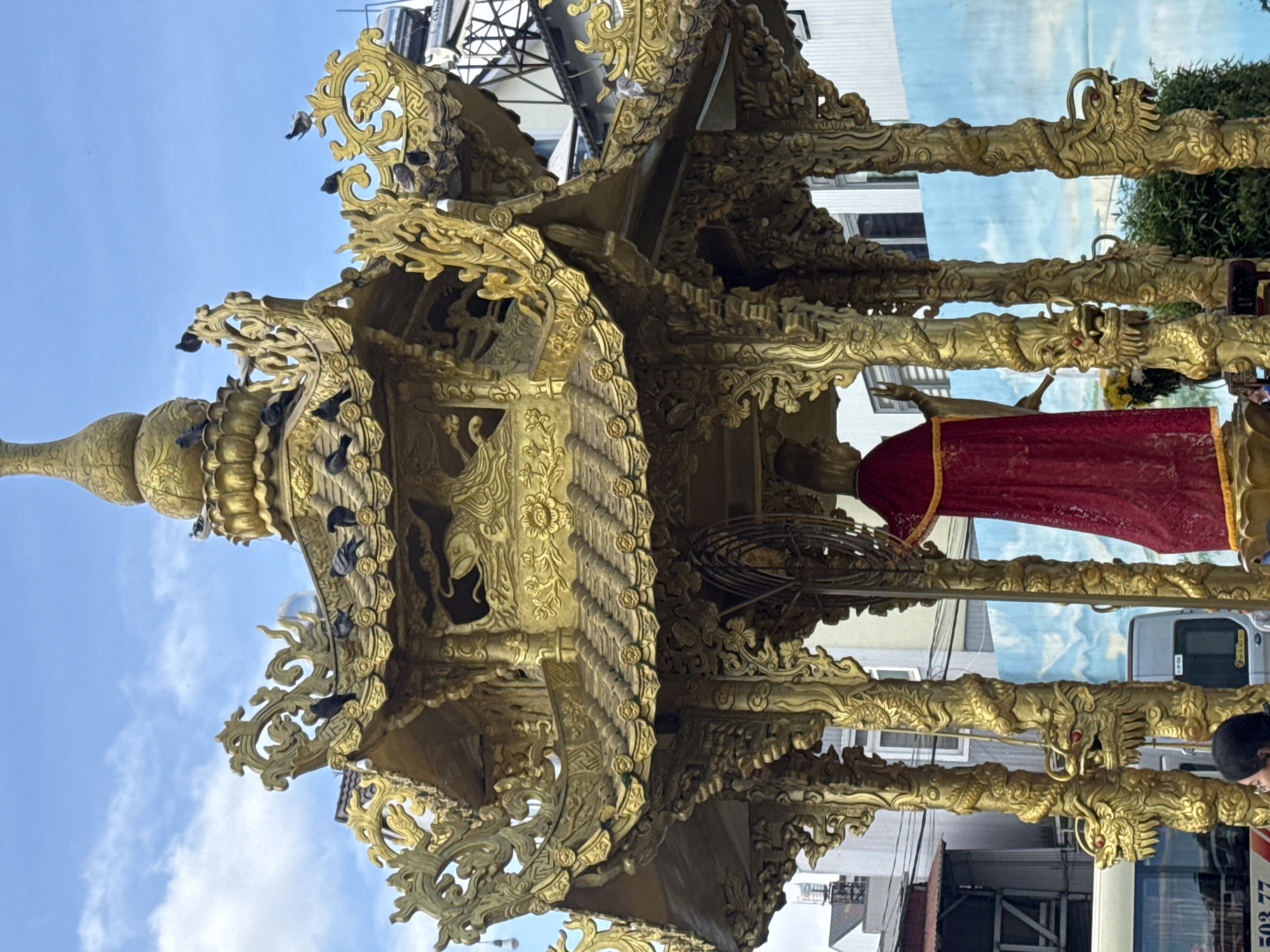 This image depicts a large, ornate golden pavilion or shrine, likely a movable float, situated outdoors in Xuân Thành, Vietnam, under a bright blue sky with scattered white clouds. The pavilion features multiple tiers of intricately carved golden roof sections and eaves, adorned with elaborate scrollwork and decorative motifs, including what appears to be a fish or sea creature carving on the upper section. Numerous dark-feathered pigeons are perched on the golden roof and its decorative elements.
The pavilion is supported by several golden columns, each intricately carved with a winding dragon motif; at the base of some columns, detailed dragon heads with prominent red eyes are visible. Inside the pavilion, a statue with golden skin or material is visible, draped in a long, deep red robe. The statue's right arm is raised, with its hand extended in a gesture.
In the background, urban elements include several buildings; one prominent building is light-colored with visible windows, and another structure features metal scaffolding and a water tank on its roof. A painted mural, predominantly light blue, depicting a landscape with mountains and water, serves as a backdrop behind the pavilion. Portions of vehicles, specifically a white bus in the lower left and a grey van, are partially visible at street level. Green foliage is present on the right side of the pavilion's base. The top of a person's head with dark hair is visible in the lower left corner of the frame. The scene is illuminated by clear daylight.