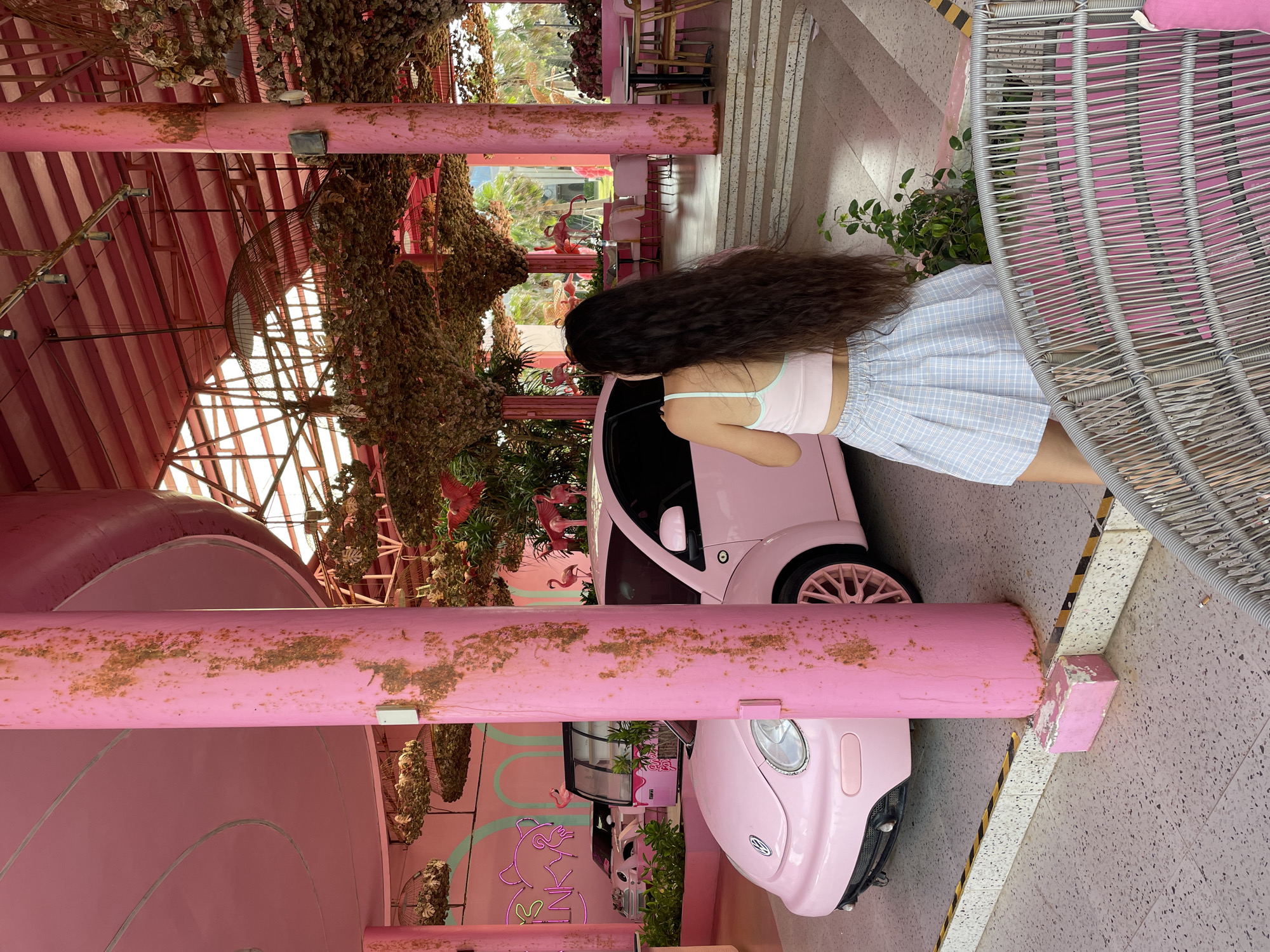 A woman with long, dark hair is standing in front of a pink Volkswagen Beetle. She is wearing a pink crop top and a light blue plaid skirt. The Beetle is parked in what appears to be an outdoor cafe or restaurant, decorated in a predominantly pink theme. There are other pink cars and a refrigerator display case visible in the background, along with neon pink signage that reads "PINKY". The ceiling is covered with decorative foliage, and there are pink flamingo statues interspersed throughout the area. The floor is a speckled terrazzo. The location is identified as a cafe in Tien Thanh Ward, Vietnam.