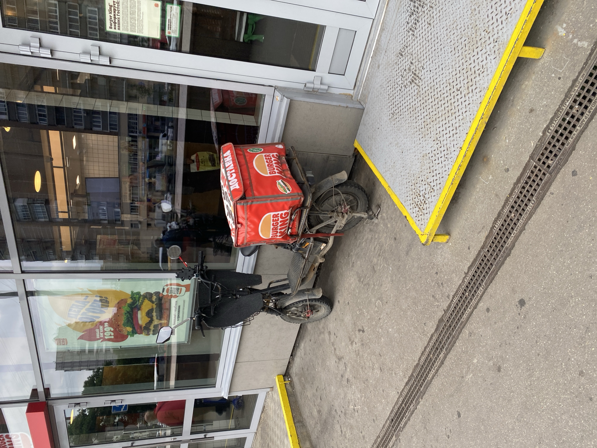 The image captures an outdoor scene in front of a Burger King restaurant in Khimki, Russia. A red electric scooter with a large red insulated delivery box is parked on the concrete pavement. The box is emblazoned with "BURGER KING" logos and the Russian word "ДОСТАВКА" (Delivery). The scooter has a black basket on the handlebars. A metal ramp with yellow trim is visible to the right of the scooter. The restaurant's glass facade is behind the scooter, reflecting the buildings across the street. A poster advertising a burger with "199" is displayed on the window. Two individuals are partially visible through the glass, one wearing a red jacket and another in darker clothing. A man in a blue shirt stands near the entrance on the right. The ground features a drainage grate.