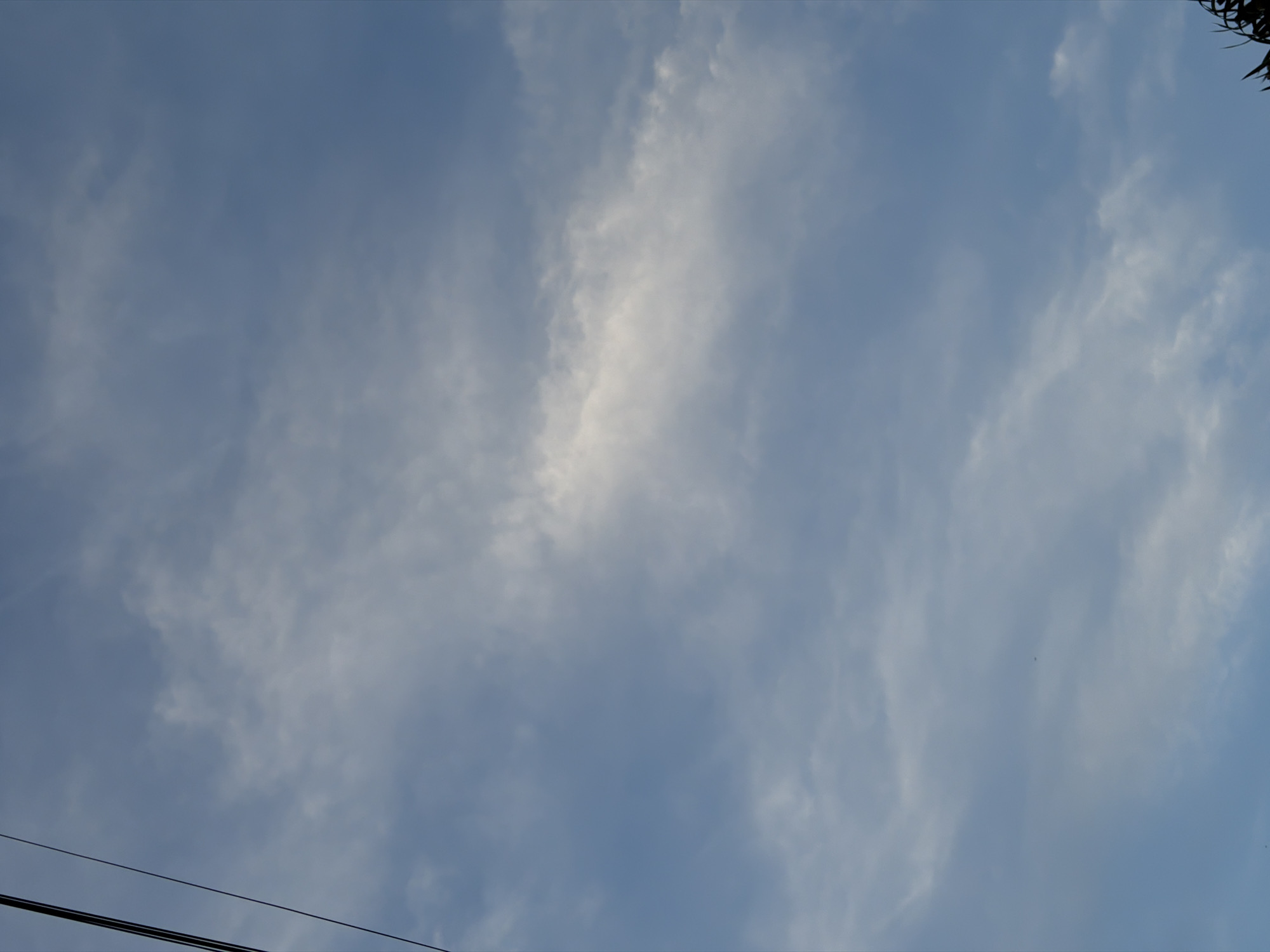 The image shows a daytime sky in North Kuta, Indonesia. The sky is light blue with scattered cirrus clouds. Power lines are visible in the lower left corner of the image. Foliage is visible in the upper right corner.