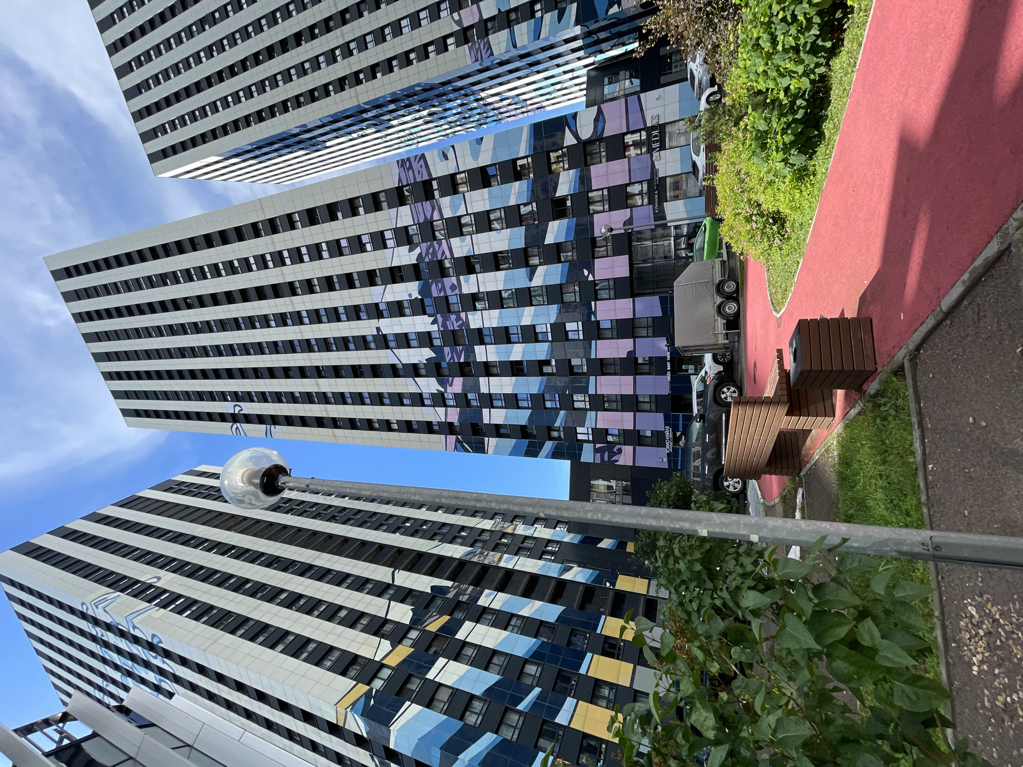 This media file depicts an outdoor scene with modern architecture, a red pathway, and lush greenery. The primary subjects are several tall, multi-story buildings with distinctive facades. One building features a white and black striped design, while another is adorned with a vibrant abstract mural in shades of blue, purple, and yellow. The buildings are tall and appear to be apartment complexes or office buildings, reflecting a contemporary urban environment.
In the foreground, a bright red, paved pathway curves through the scene. Adjacent to the pathway is a wooden bench and planter. To the right of the pathway, a patch of dense, green bushes and trees adds a natural element. In the background, several vehicles are visible, including a white car, a dark SUV, a green car, and a truck with a trailer, suggesting a residential or commercial area. The sky is a clear blue with a few wispy clouds, indicating fair weather.
There is no discernible activity or event taking place; the scene appears to be a still, environmental shot. The time of day seems to be daytime, likely late morning or early afternoon, given the lighting and the shadows cast. The location cues, such as the architecture and the Cyrillic text visible on one of the buildings ("КЛИНИКА" which means "CLINIC"), point to Moscow, Russia. The overall mood is calm and orderly, showcasing a blend of modern urban design and natural elements.