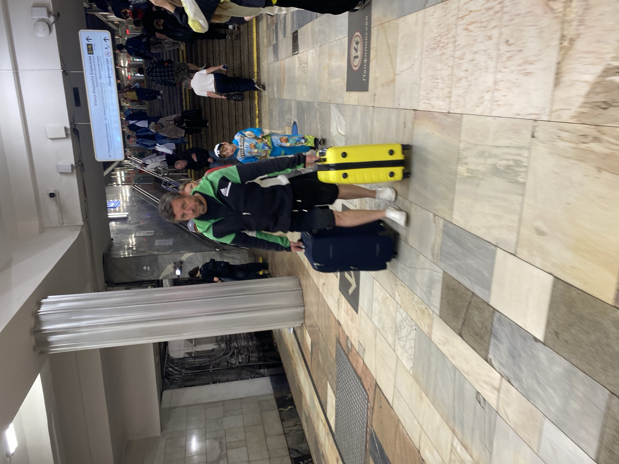In an indoor public transit station, likely a metro station, a man and a young boy are walking on a marble-tiled floor. The man, dressed in a black and green athletic jacket and black shorts, is pulling a dark blue suitcase. Beside him, the boy, wearing a blue jumpsuit with cartoon characters, is pulling a bright yellow suitcase. Both are walking towards the viewer. In the background, other commuters are seen walking up a set of stairs, with a directional sign overhead indicating "Ulitsa Marshala Biryuzova" and "Panfilovskaya". The environment appears clean and well-lit, consistent with a modern transit hub. The location is identified as the Khimki, Russia metro station.