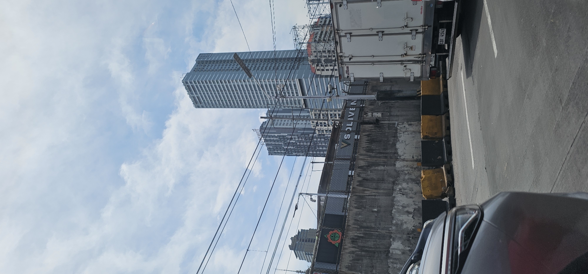 The image captures a street-level view of Quezon City, Philippines, on a partly cloudy day. In the foreground, the front left portion of a dark-colored car is visible on the right side of the frame. A white truck with the brand "ISUZU" visible on its rear is positioned in the right lane, facing away from the viewer. The truck's license plate reads "CDF 4143". A concrete barrier, painted with alternating yellow and black sections, lines the road.
Behind the barrier, a textured concrete wall is prominent, featuring a metallic fence at its top. A circular logo with orange and green elements is visible on the left side of the fence, and a sign with illuminated letters reads "VS SOLIVEN".
In the background, several high-rise buildings dominate the skyline. The tallest and most prominent building is a light blue skyscraper with a grid-like facade, labeled "GRAND TOWER" and "TRIBE". To its left is a shorter, dark gray skyscraper with a similar grid pattern, bearing the sign "SUMMIT". Further in the distance, other buildings with varied architectural styles are visible, along with electrical wires crisscrossing the sky. The sky itself is a light blue, interspersed with scattered white clouds.