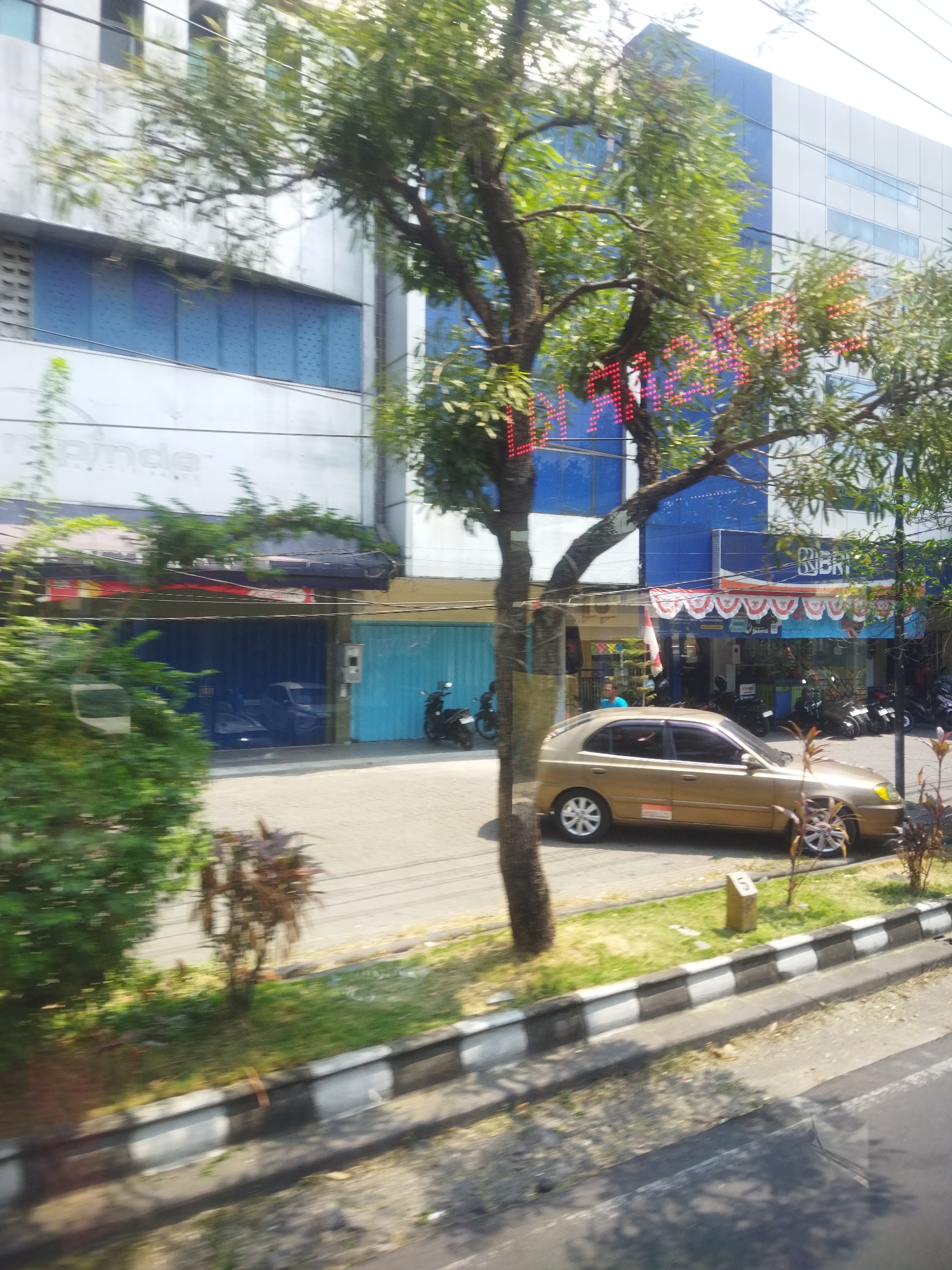 The image depicts a street scene in Surakarta, Indonesia, on a sunny day. In the foreground, a golden sedan is parked on the side of the road, facing right. Behind the car, a sidewalk with a black and white striped curb runs along the street. Several motorcycles are parked near the buildings. The buildings lining the street appear to be a mix of commercial establishments. One building has a modern facade with blue and white panels, and a digital display with red and pink lights that seems to form text. Another building has a more traditional awning with red and white accents. Trees with green foliage are present along the street, casting shadows on the ground. The overall impression is a busy urban environment with a mix of modern and traditional architecture.