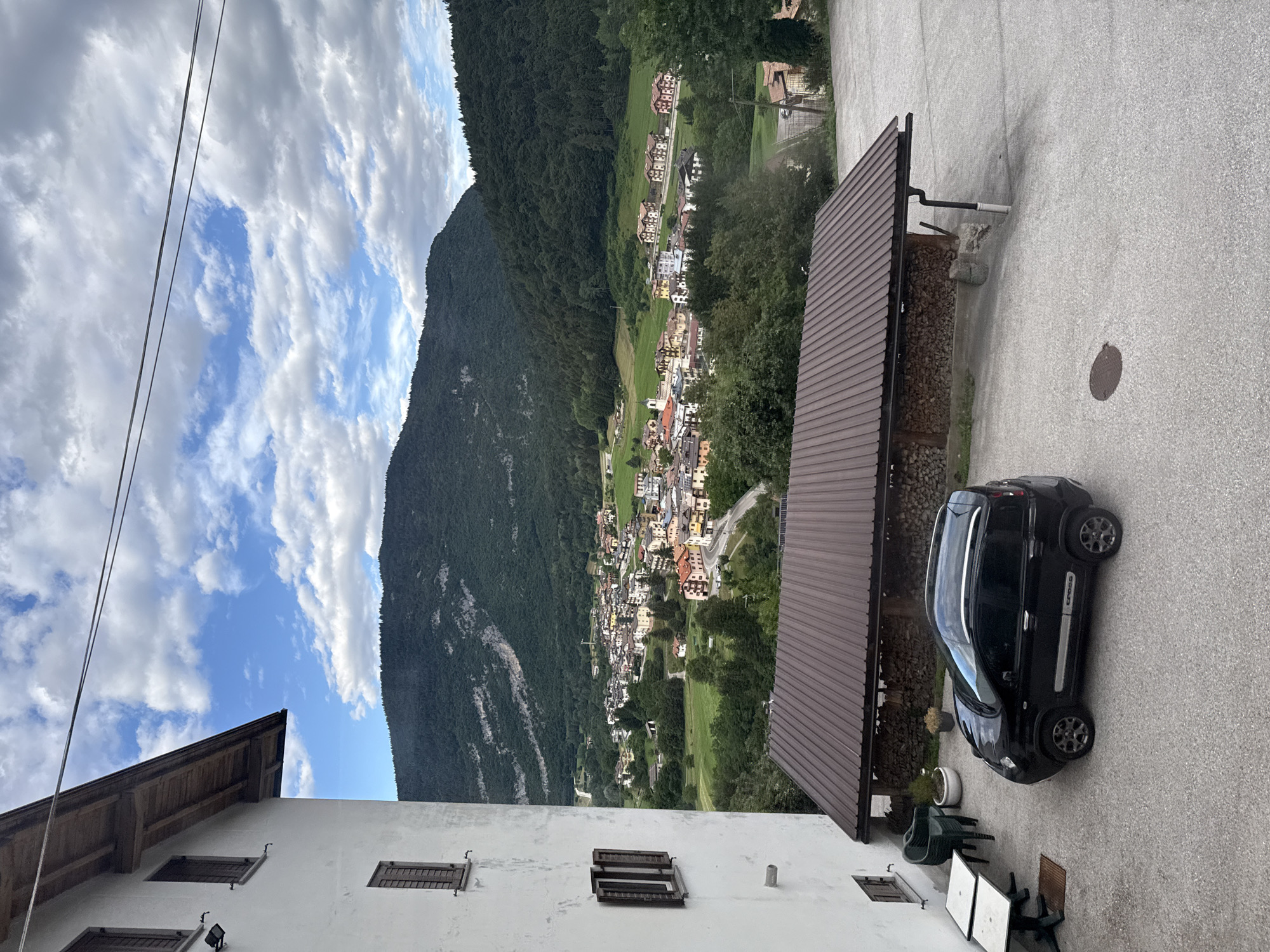 This is a photograph taken from an elevated perspective, looking down onto a small town nestled in a valley at the base of a lush, tree-covered mountain. The sky is partly cloudy, with patches of blue visible between white, fluffy clouds. The sun appears to be shining, suggesting it is daytime.
In the foreground, the edge of a white building with wooden window shutters is visible. To the right of this building, a dark-colored car is parked on an asphalt surface. Adjacent to the car is a covered structure with a brown, corrugated roof, and stacks of firewood underneath. Beyond the car and the covered structure, the town unfolds in the valley. The buildings are a mix of colors, including white, yellow, and red, and they are clustered along winding roads. The town is surrounded by green fields and trees.
The mountain that dominates the background is densely forested, with some patches of rock visible on its slopes. The overall scene depicts a tranquil, rural setting in a mountainous region. The time of day appears to be afternoon, given the angle of the sunlight. There is no visible text in the image. The location context provided is Cavedago, Italy, which aligns with the European architectural style of the buildings and the mountainous landscape.