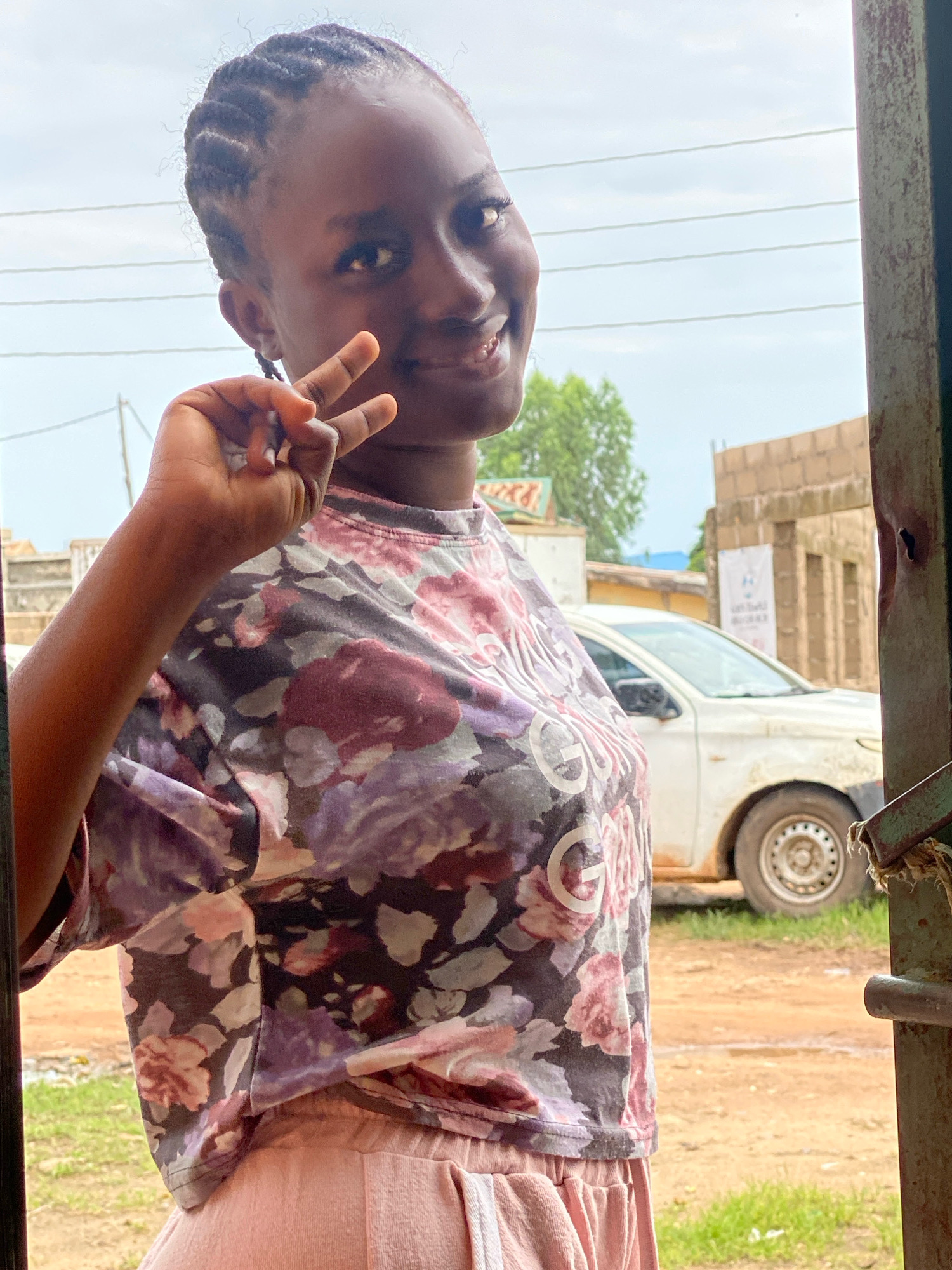 A young Black woman with braided hair and a floral-patterned shirt smiles and holds up two fingers in a peace sign. She is standing in front of a dirt road in Sabon Tasha, Nigeria. A white car is parked behind her, and there are buildings and trees in the background. The scene appears to be outdoors, possibly near a market or residential area, with an overcast sky.