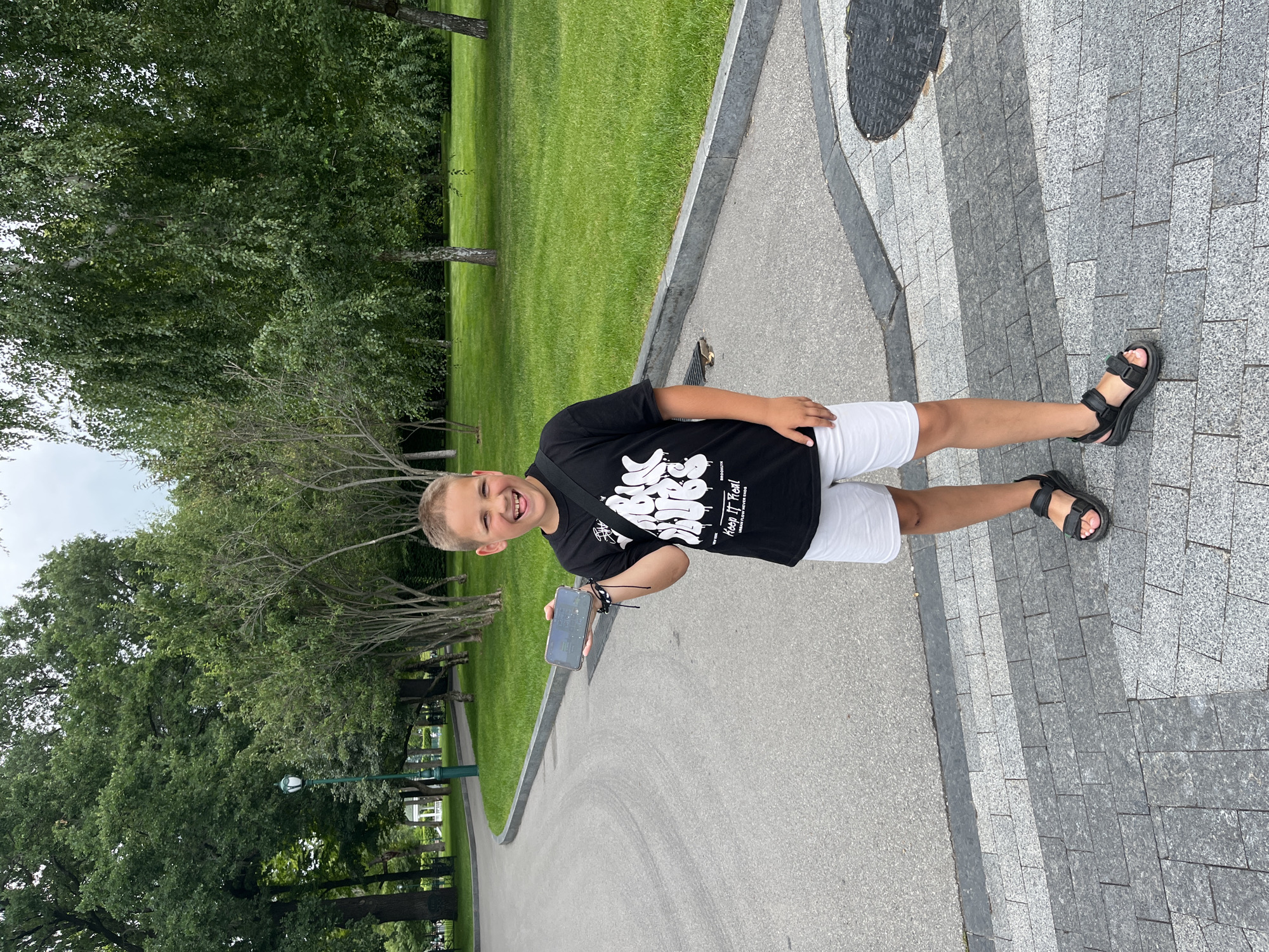 In a park in Kharkiv, Ukraine, a young boy with fair hair stands on a paved walkway, holding a mobile phone in his right hand. He is wearing a black t-shirt with white graffiti-style lettering and white shorts, along with black sandals. The boy is smiling broadly, with his tongue sticking out slightly. Behind him, a path curves through a green lawn bordered by trees. The sky is overcast.