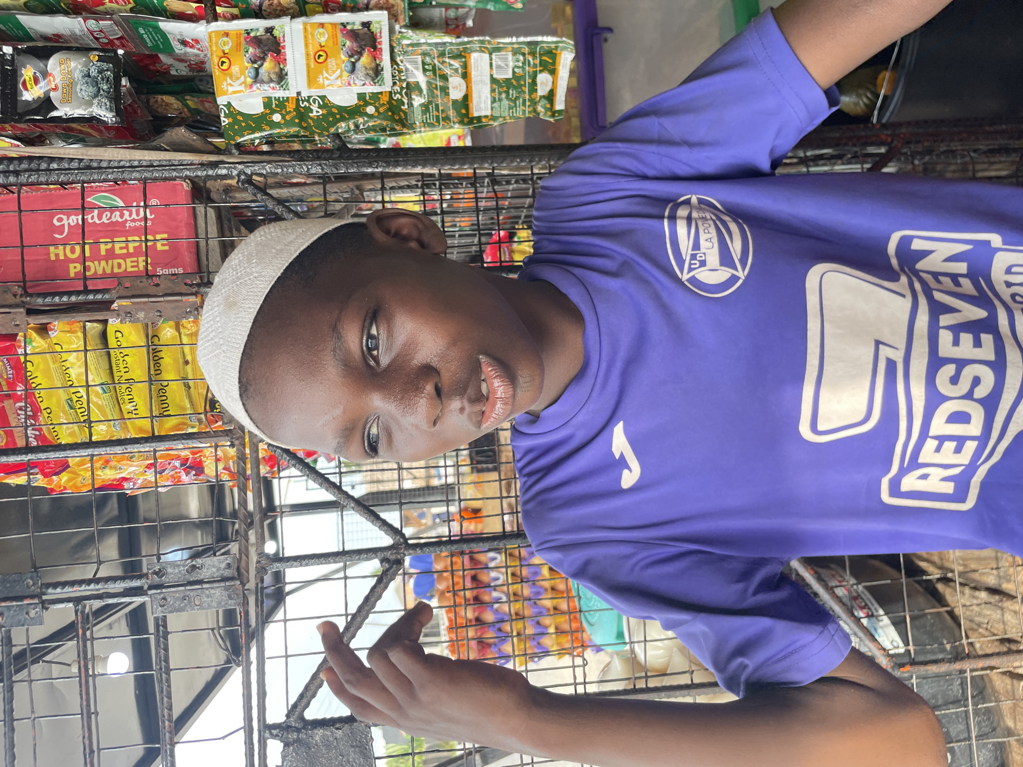 A young Black person, likely a boy, is depicted in a close-up, sideways shot within an outdoor market or kiosk setting in Badagry, Nigeria. He wears a white kufi cap and a purple t-shirt with visible white logos, including "U.D. LA POVEDA" and "REDSEVEN." His gaze is directed towards the camera with a neutral to slightly curious expression.
The background is filled with various packaged food items displayed on metal grid shelves. Products include "Golden Penny Instant Noodles," "godeart foods HOT PEPPE POWDER 5ams," "Dawa Dawa Seasoning Powder," "Tomato Mix," and other "SPICES." Several packets feature illustrations of chickens or roosters. The metal grid structure suggests a typical street vendor stall or small shop. The lighting indicates it is daytime. The overall scene captures a moment from daily life and commerce in a local Nigerian market.
