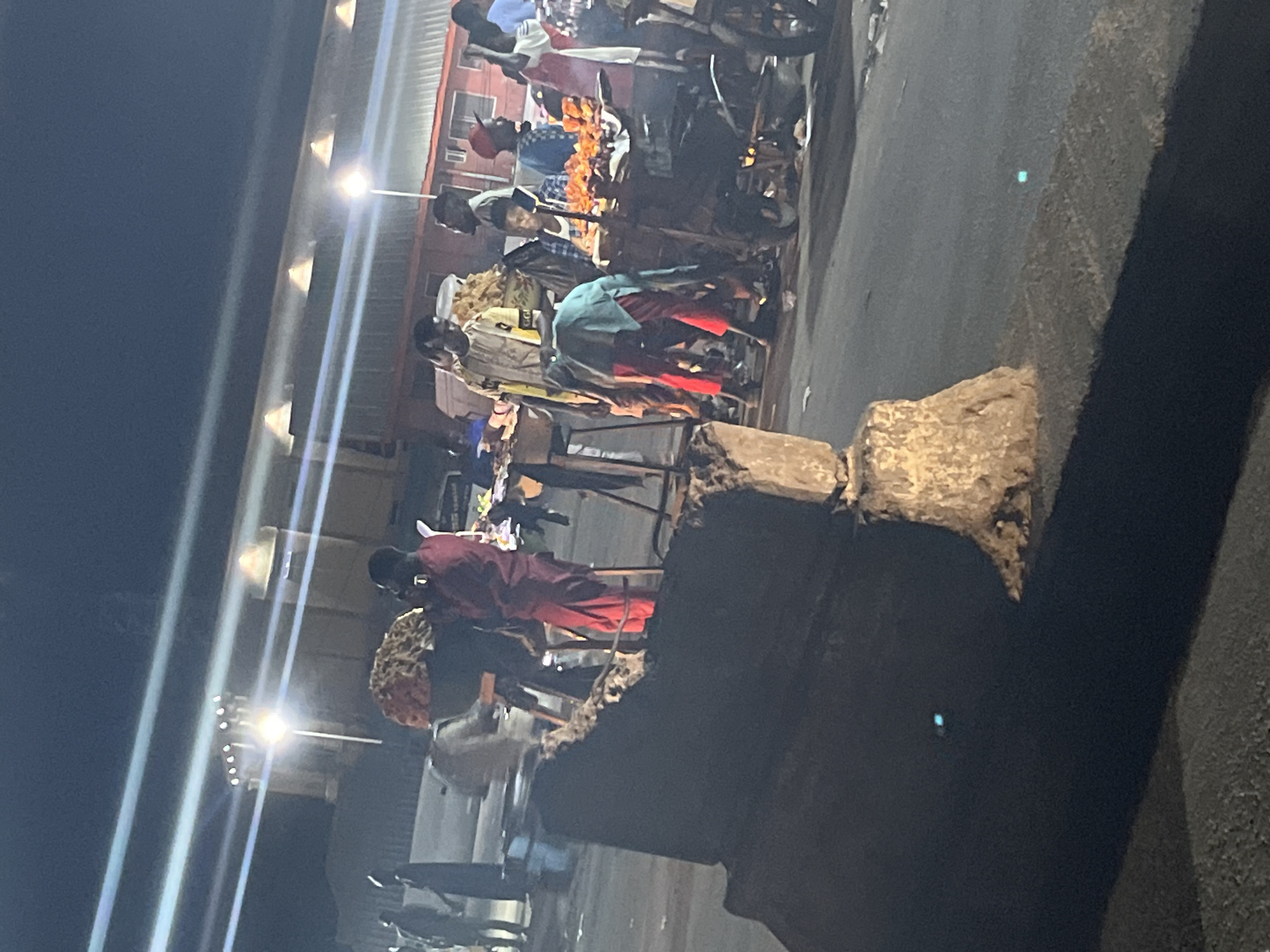 This is a nighttime outdoor scene at a market stall in an urban environment. The lighting is artificial, with bright overhead lights casting long shadows. A few individuals are visible, some appearing to be vendors and others customers, engaged in activities related to the market. One person in the foreground on the left is partially obscured by a large concrete structure. In the background, there are buildings with illuminated windows and a row of lights across the top of the structure, suggesting it might be a covered market or a building facade. The ground appears to be paved asphalt. There are several market stalls with food displayed, possibly grilled meats or other prepared food items. The overall impression is a bustling, albeit dimly lit, night market.