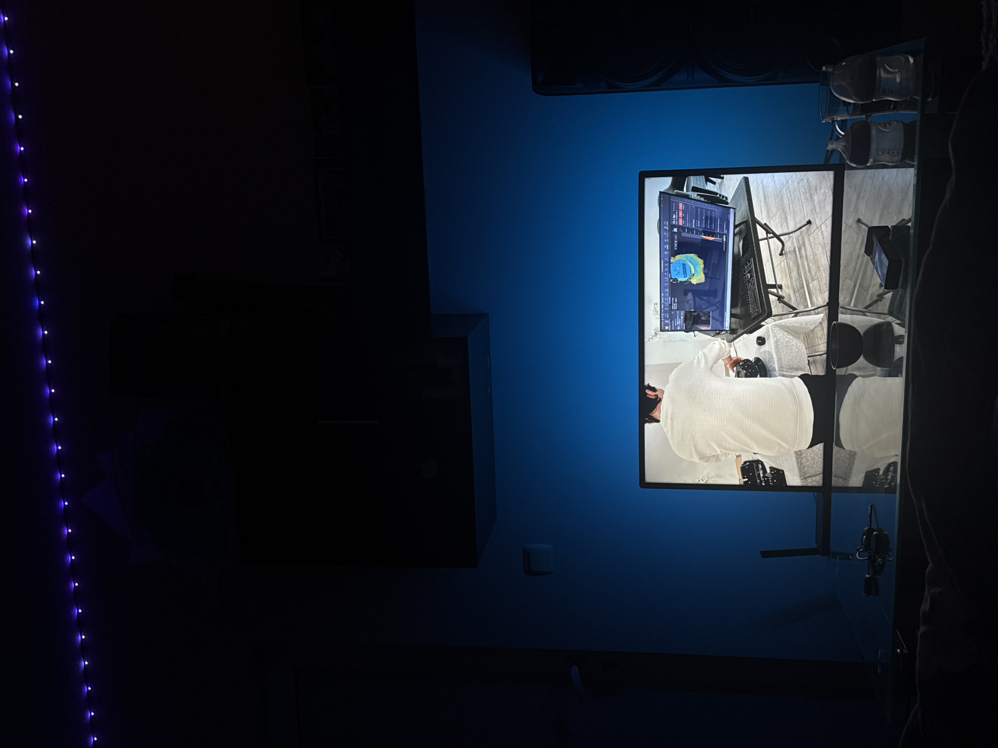 Here's a detailed description of the image:
**Scene:** The image shows an interior setting with a focus on a wall-mounted multi-screen display. The environment has a modern feel, with dark furniture and blue-toned lighting.
**Objects:**
* A multi-screen display showing various video feeds. One screen seems to show a person (in white clothing), and another features a display with data visuals and controls.
* A dark cabinet or storage unit is visible to the left of the screen.
* A string of purple LED lights runs vertically along the left edge of the frame.
* Other objects are visible in the video feeds on the screens, including a laptop, headphones, and camera equipment.
**Environment:**
* The room is dimly lit, primarily illuminated by the blue lighting and the screens.
* The walls are painted blue.
**Location:** It is not possible to determine if the location is in Sofia, Bulgaria without further information.