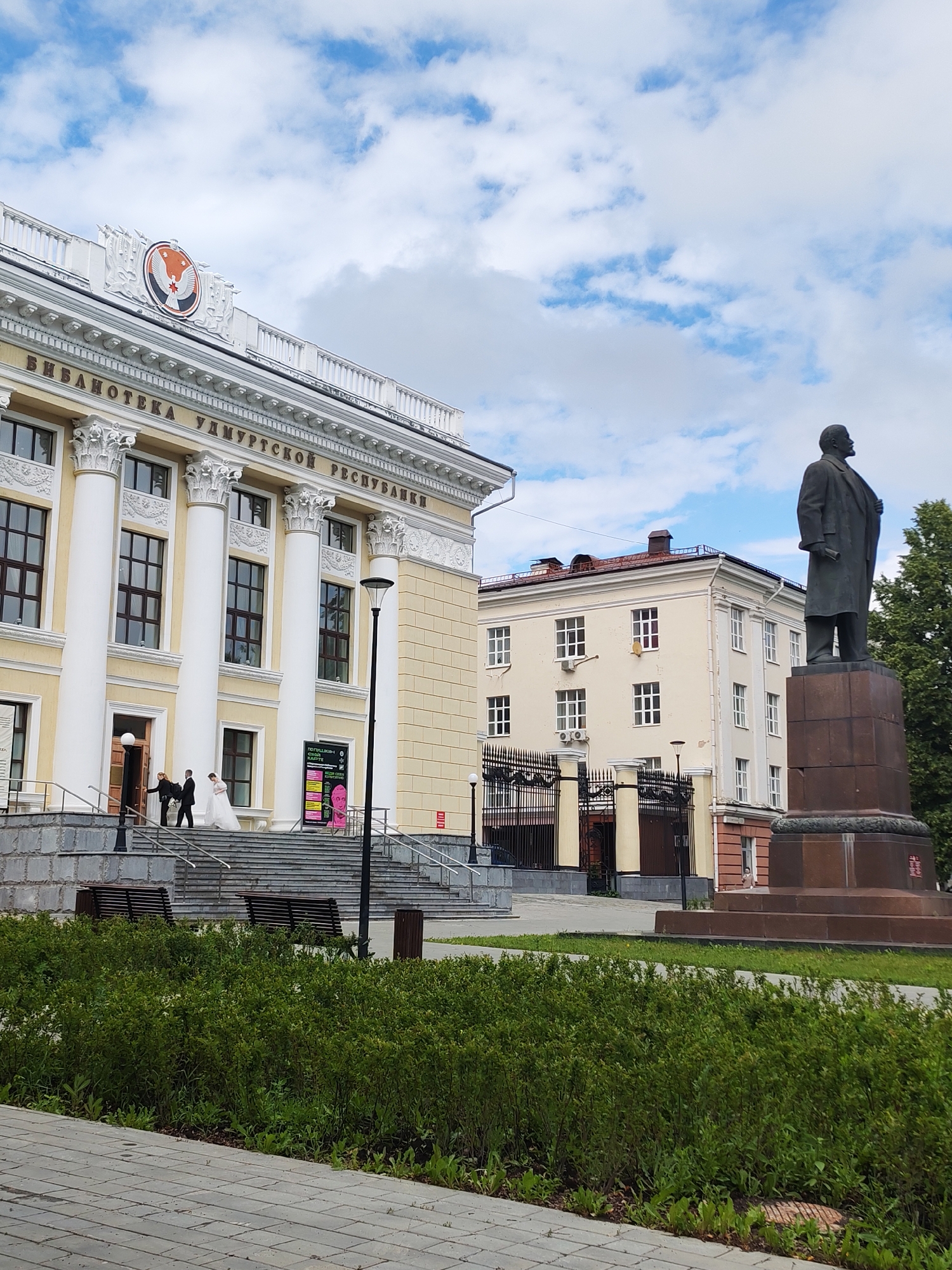 In Izhevsk, Russia, on a cloudy day, a wedding party is captured on the steps of a grand, yellow building. The building, adorned with classical columns and a decorative frieze, bears the inscription "БИБЛИОТЕКА УДМУРТСКОЙ РЕСПУБЛИКИ" (Library of the Udmurt Republic). A bride in a white gown and a groom in a dark suit are ascending the stairs, accompanied by another person. To the right of the building, a tall statue of Vladimir Lenin stands on a pedestal, gazing into the distance. In the foreground, lush green bushes and paved walkways create a park-like setting. The overall scene depicts a blend of civic architecture, historical monument, and personal celebration.