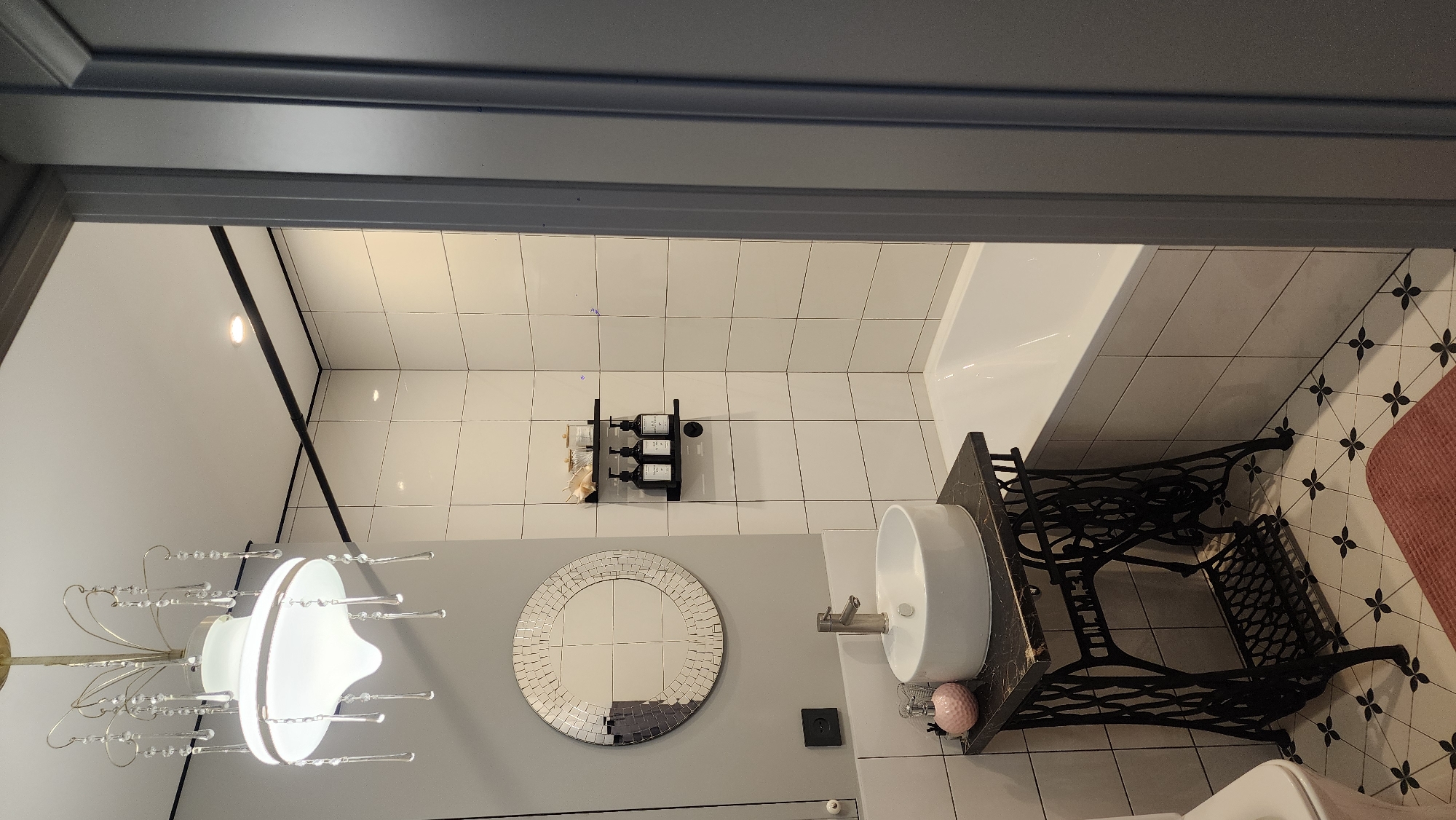 The image shows a bathroom interior. A white circular sink sits atop a black wrought-iron stand, reminiscent of an old sewing machine base. A round, mosaic-bordered mirror hangs on the gray wall above the sink. To the left, a chandelier with decorative crystals is visible. Above the sink, on a tiled wall, a black shelf holds toiletries. A white bathtub is situated to the right. The floor has patterned tiles. A partial doorway frames the scene. There is no indication of this being in Smolensk, Russia.