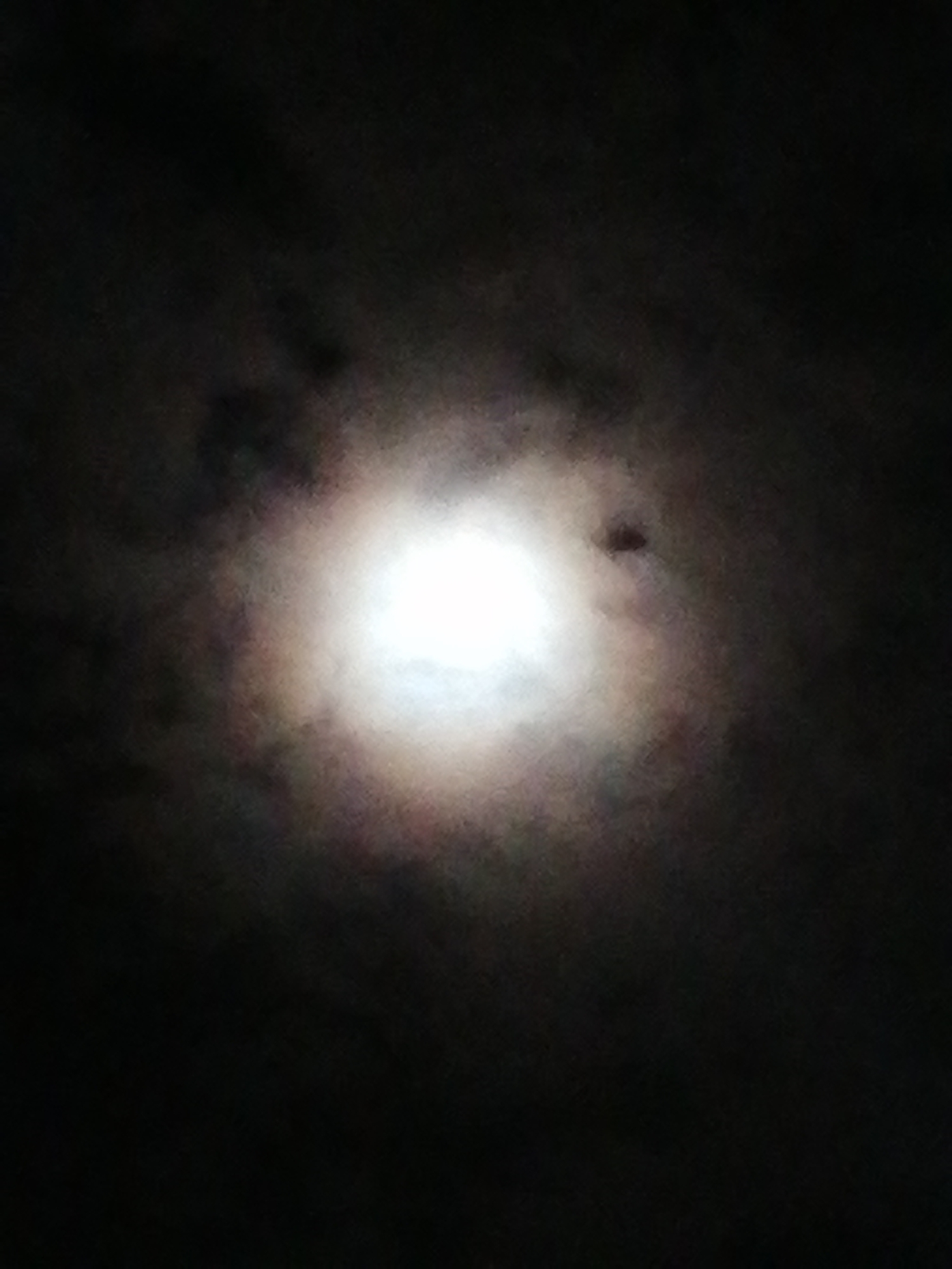 The image depicts a bright light source, likely the moon, shining through a dark, cloudy sky. The light is diffused by the clouds, creating a soft, blurred glow. No people or identifiable objects are visible besides the light source and clouds. There is no information to infer any specific location in Da Lat, Vietnam.
