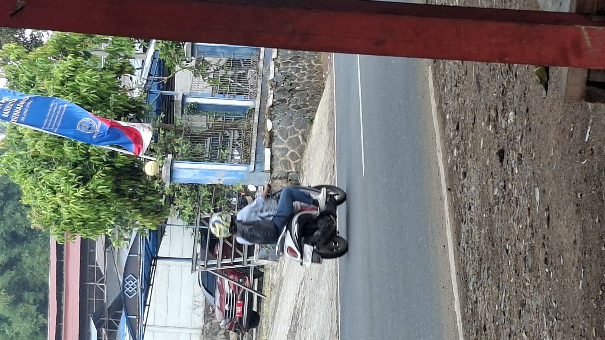 A medium shot captures a person riding a scooter on a paved road in Cilaku, Indonesia. The rider, wearing a blue long-sleeved shirt, blue jeans, white sneakers, and a yellow and blue helmet, is seen from the rear and right side, carrying a black backpack. The scooter is white with black accents and is in motion. In the background, a red car is parked on the left, and a blue and white building with a metal fence and a stone wall are visible. A blue and red flag with text in Indonesian is fluttering on a pole to the right of the building, near some green trees. The road surface has a white line marking its edge. The foreground shows a gravelly shoulder with some debris.