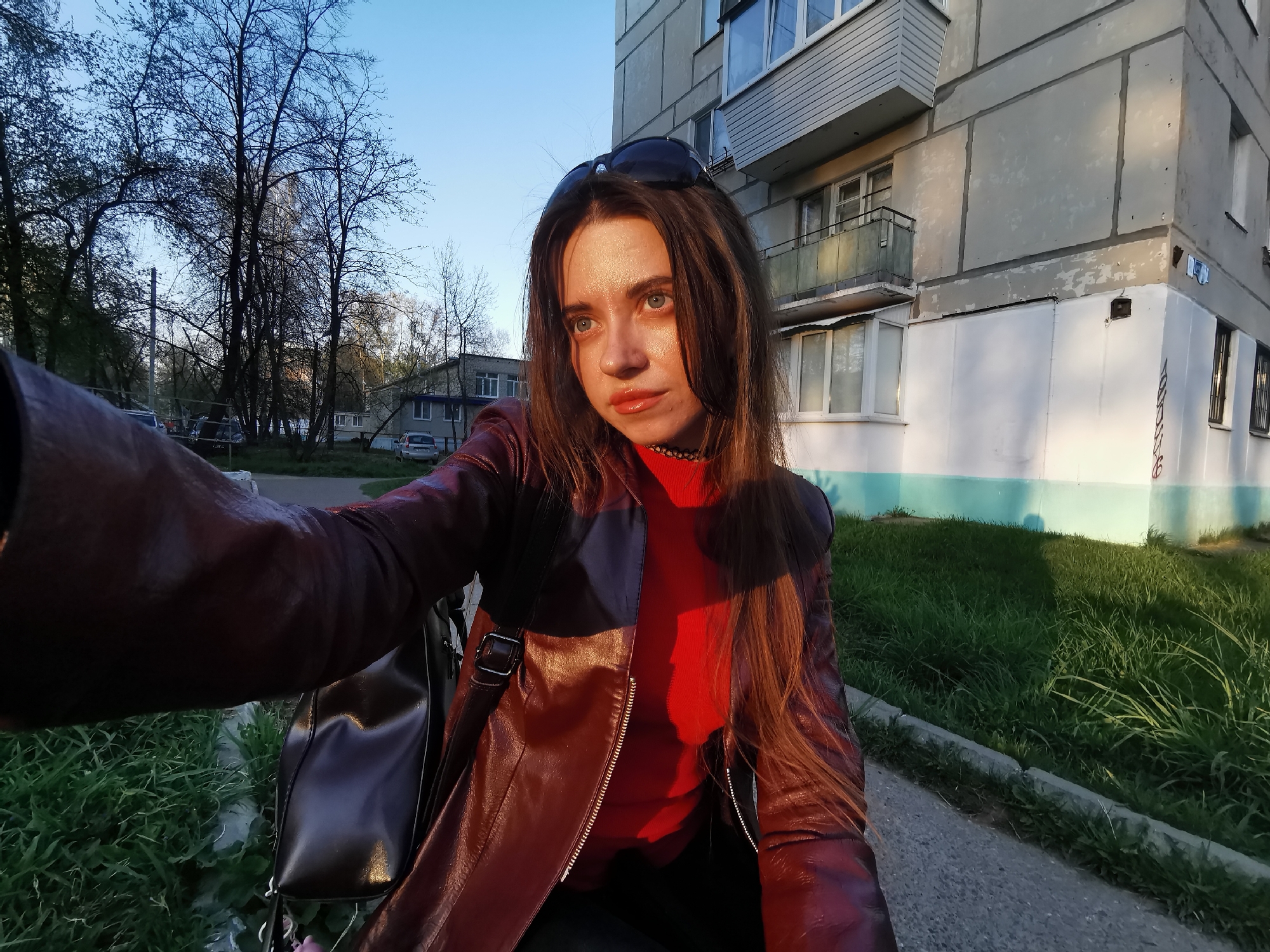 A young woman is taking a selfie outdoors. She has long brown hair and is wearing sunglasses on her head. She is dressed in a red top and a reddish-brown leather jacket. A black backpack is slung over her shoulder.
The background shows a multi-story building with a weathered, gray and white exterior. The building features windows and small balconies. To the left, there are bare trees and a glimpse of another building in the distance.
The immediate foreground consists of a paved walkway and patches of green grass. The lighting suggests it is either early morning or late afternoon, with the sun casting shadows across the scene. The location is an urban environment, likely a residential area.
