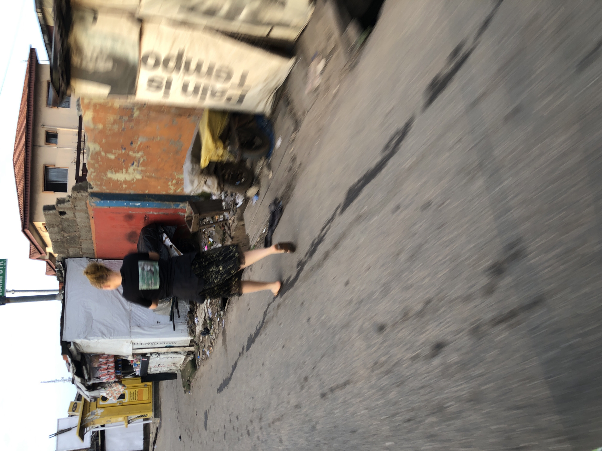The image depicts a street scene in Isheri, Nigeria, with a woman walking away from the camera on a paved road. The woman, with blonde hair pulled back, wears a black t-shirt with a rectangular graphic and a dark, patterned skirt. She is barefoot on her left foot and wearing a sandal on her right. The environment shows various structures, including a yellow kiosk with signage, makeshift stalls with white coverings, and a building with an orange and red facade. Debris and trash are visible along the side of the road and in front of the stalls. In the background, a two-story building with a red roof and windows is visible. The overall impression is of a busy, urban environment with signs of commercial activity and a somewhat unkempt appearance. The image is taken from a moving perspective, indicated by the motion blur.