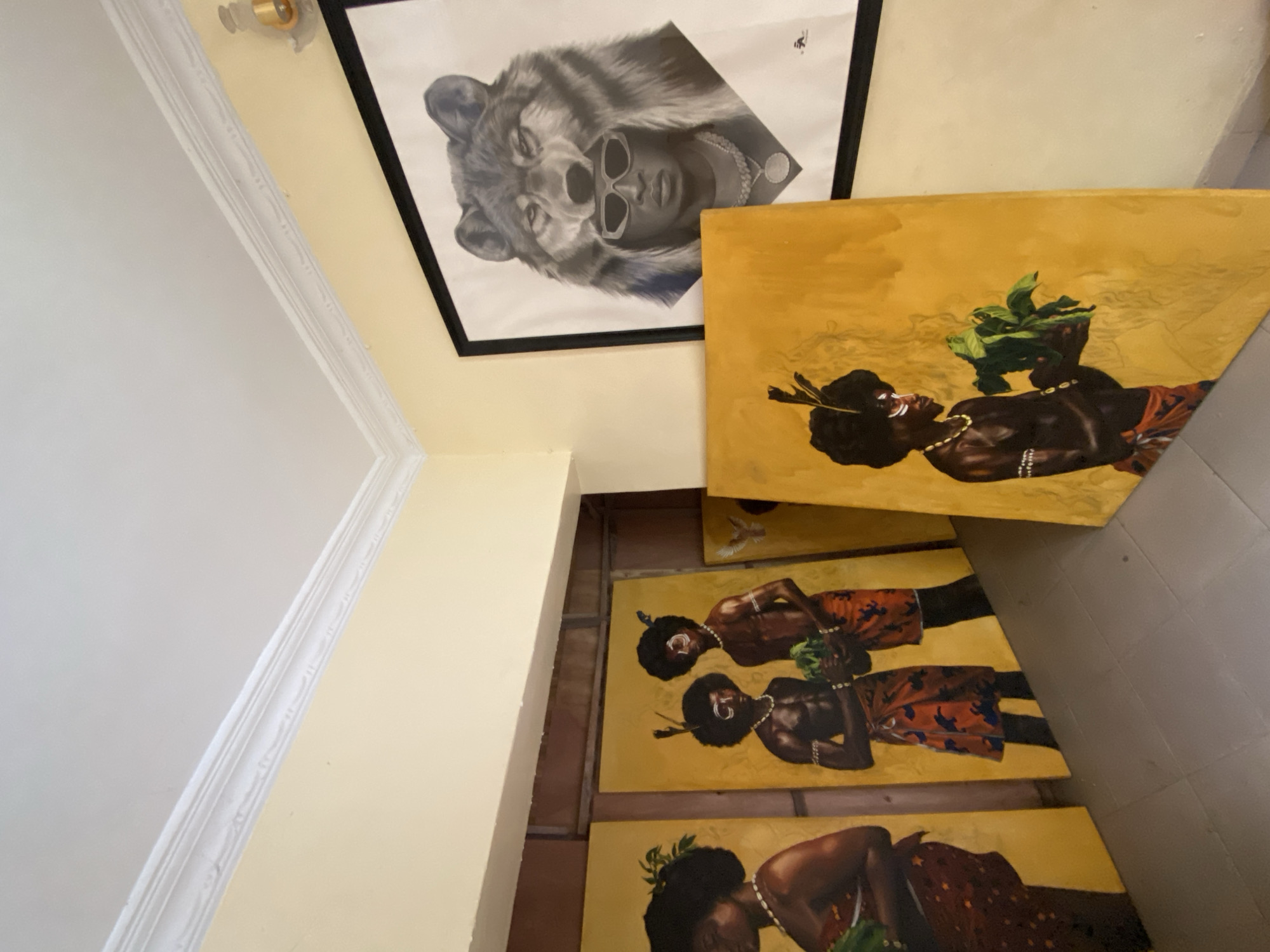 Indoor scene, likely a residence or gallery in Ado, Nigeria, featuring artwork. One framed piece is a black and white drawing of a person with a wolf head, wearing sunglasses. Adjacent to it are multiple rectangular canvases depicting black figures, possibly people of African descent, dressed in traditional clothing, set against a yellow background. The canvases appear stacked against a wall, possibly for display. The floor is tiled. Walls are painted in varying shades of yellow and white.