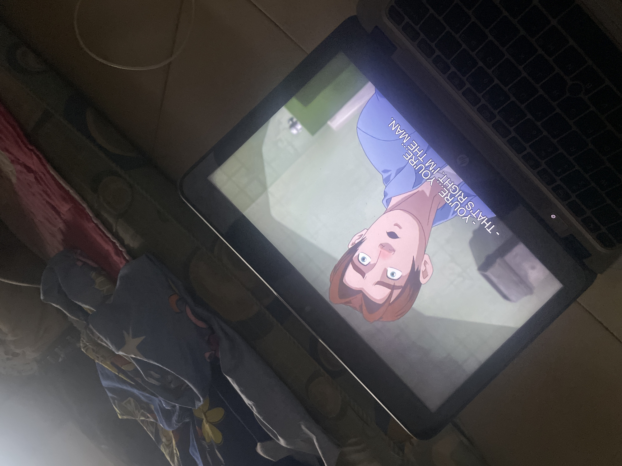 An animated male character with brown hair and blue eyes is displayed on a laptop screen, appearing surprised. He is wearing a blue shirt. Subtitles on the screen read: "- YOU'RE... YOU'RE. - THAT'S RIGHT. I'M THE MAN." The laptop is positioned on a surface that appears to be a floor, with a patterned blanket or fabric draped in the background. The environment suggests an indoor setting. The image is taken from a high angle, with the laptop screen being the focal point. The location is Ihiagwa, Nigeria.