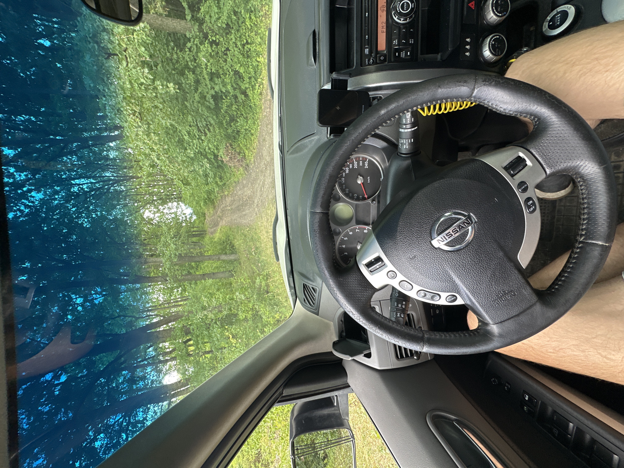 The image shows the interior of a Nissan X-Trail SUV, viewed from the driver's perspective. The car is on a dirt road surrounded by dense green forest. The dashboard, steering wheel with the Nissan logo, and driver's legs are visible. The windshield offers a view of the road ahead, which winds through trees and vegetation. The car appears to be stationary. The scene is set in Makiv, Ukraine, likely on a rural or forested road.