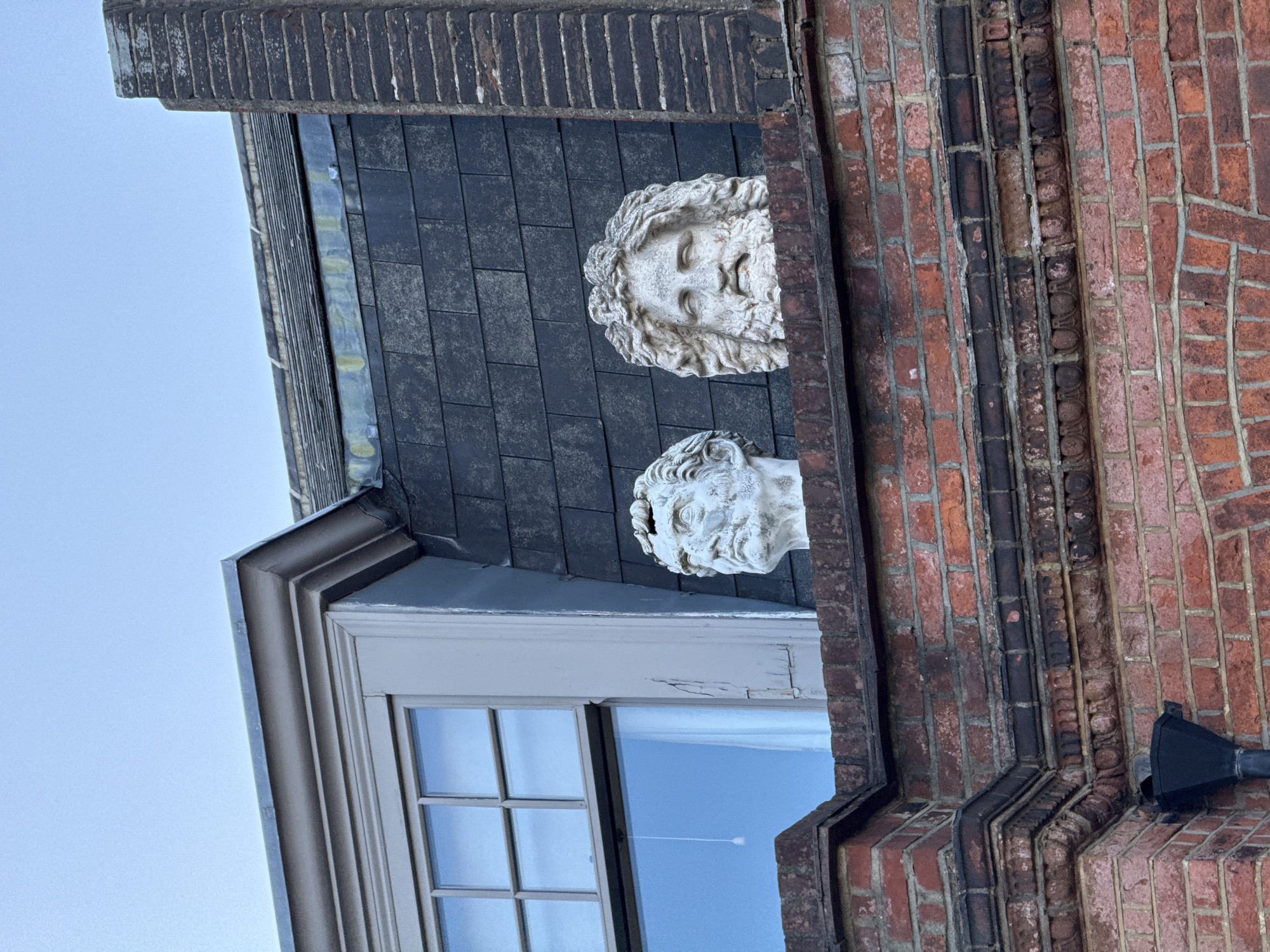 The image shows a partial view of a building in London, United Kingdom. The building exterior consists of brickwork, including red and dark bricks arranged in patterns, and slate tiles on a partial roof area. Two white, sculptural heads are mounted on the slate roof. A multi-pane window is visible below the sculptural heads. The sky is visible through the window and in the background.